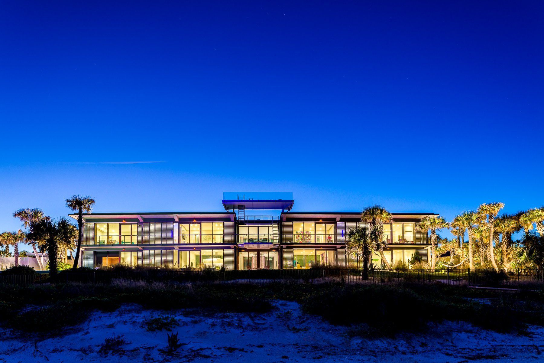 Modern, illuminated beach house at dusk, clear blue sky.