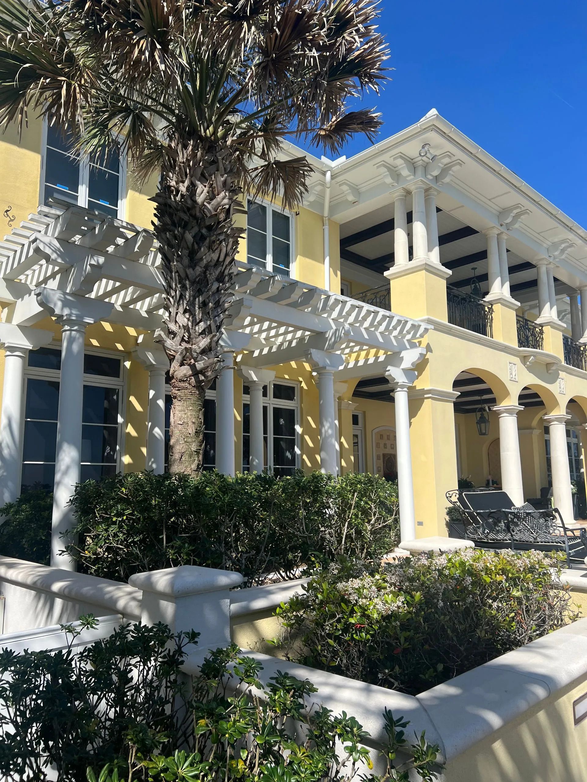Yellow historic house with white trim, arched porch, and lush shrubs in bright sunlight
