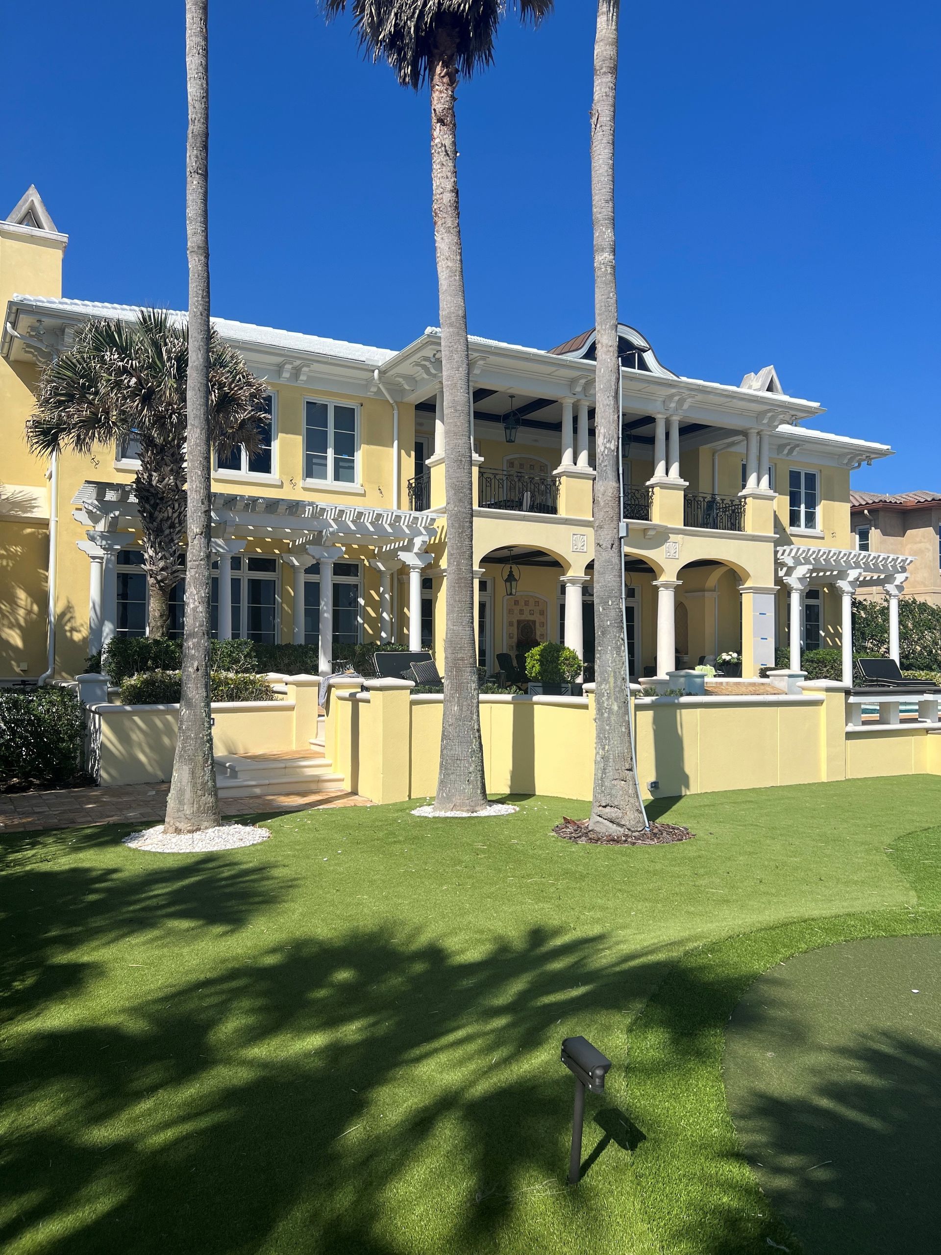 Yellow historic mansion with white trim and palm trees behind a green lawn under a clear blue sky