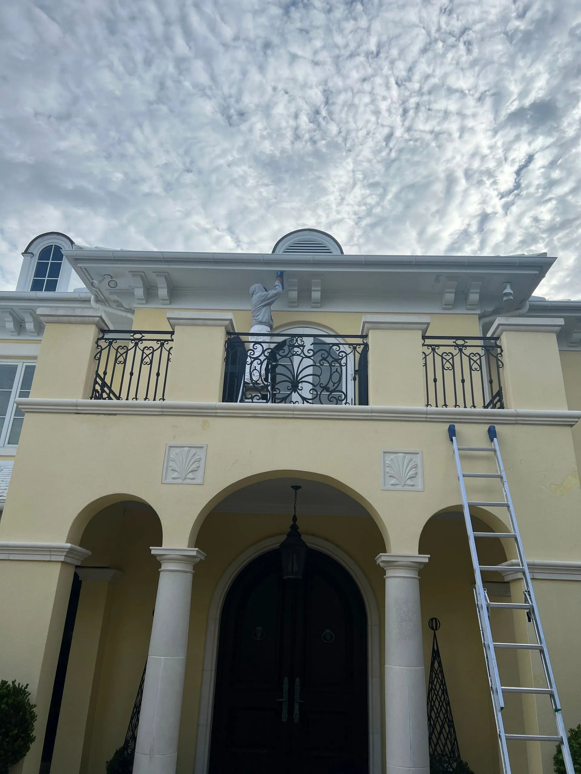 Cream-colored house facade with arched entry, balcony railing, and a ladder leaning on the right side.