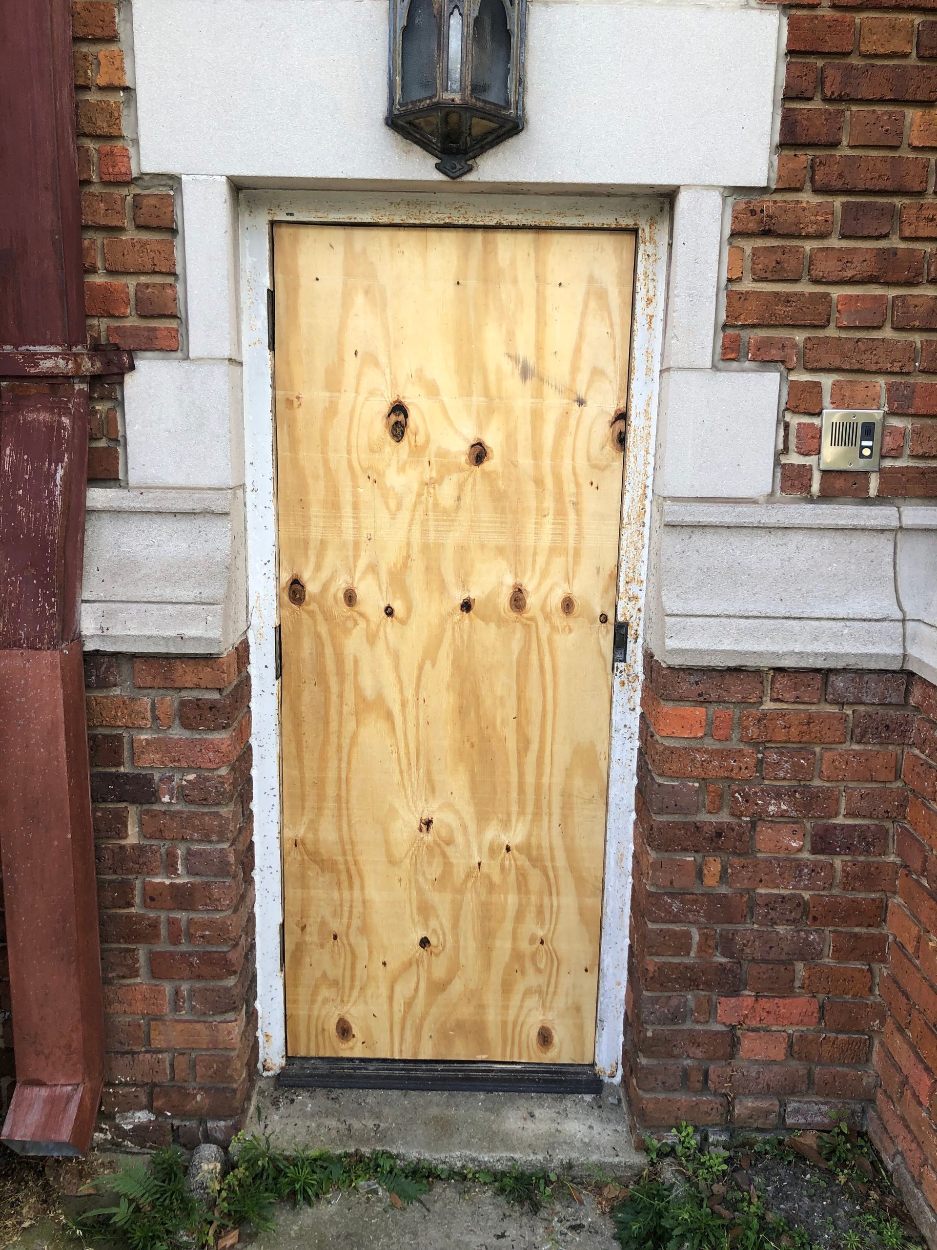 Boarded-up wooden door set in a red brick building with stone accents.  Door has visible screw holes.