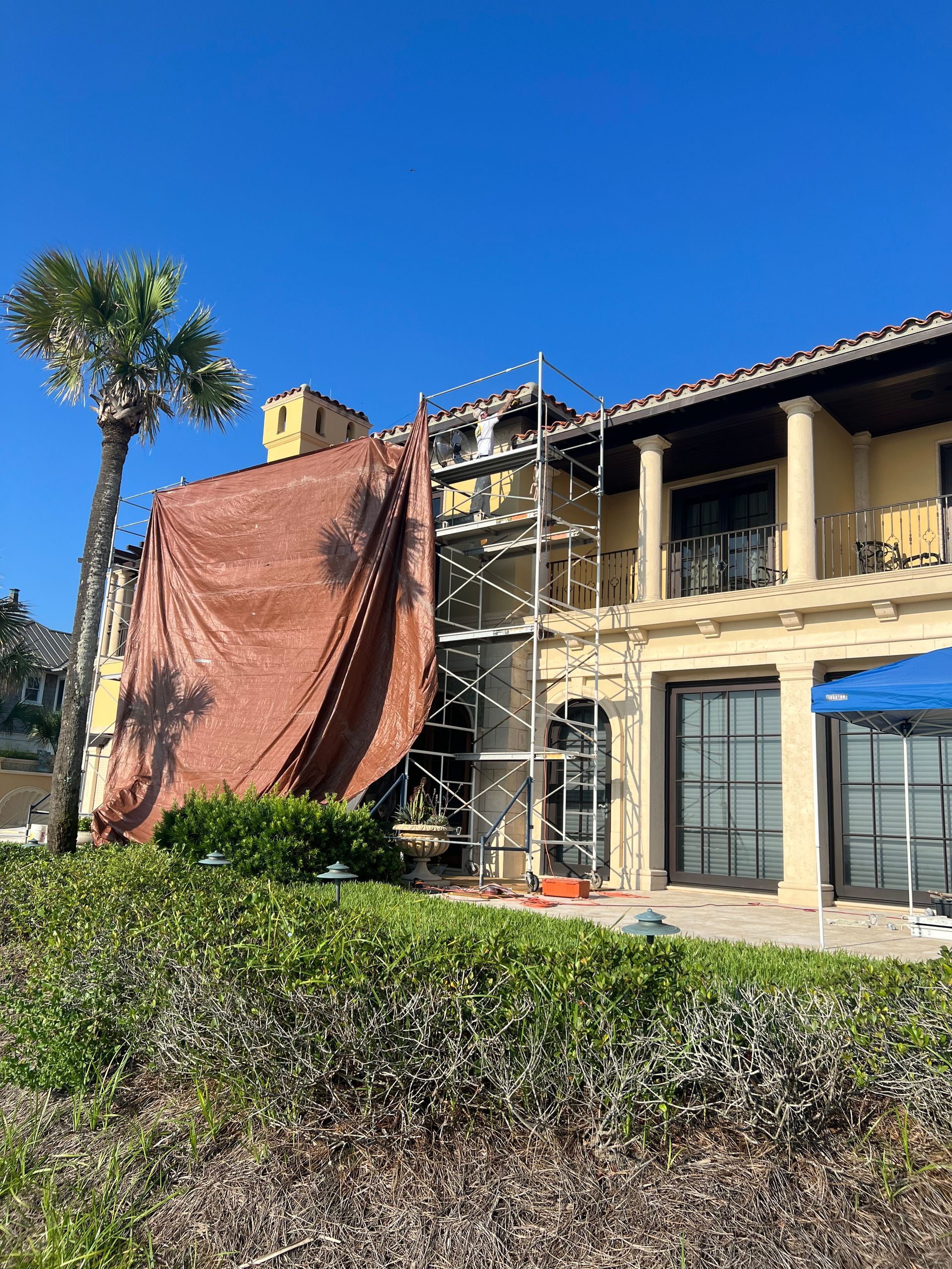 Two-story house with scaffolding, covered in brown tarp. Palm tree and clear blue sky.