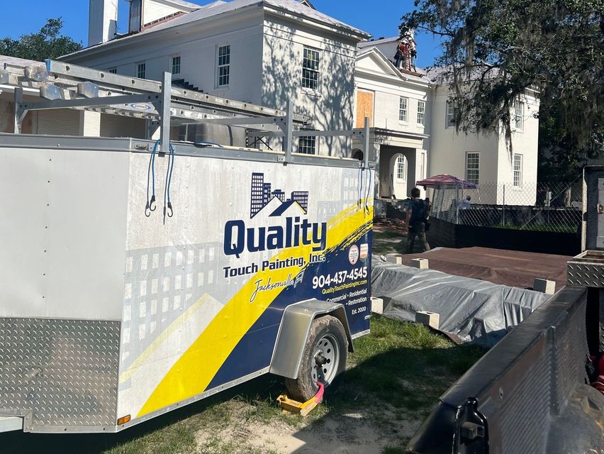 White utility trailer with “Quality” logo parked beside a house in sunlight.