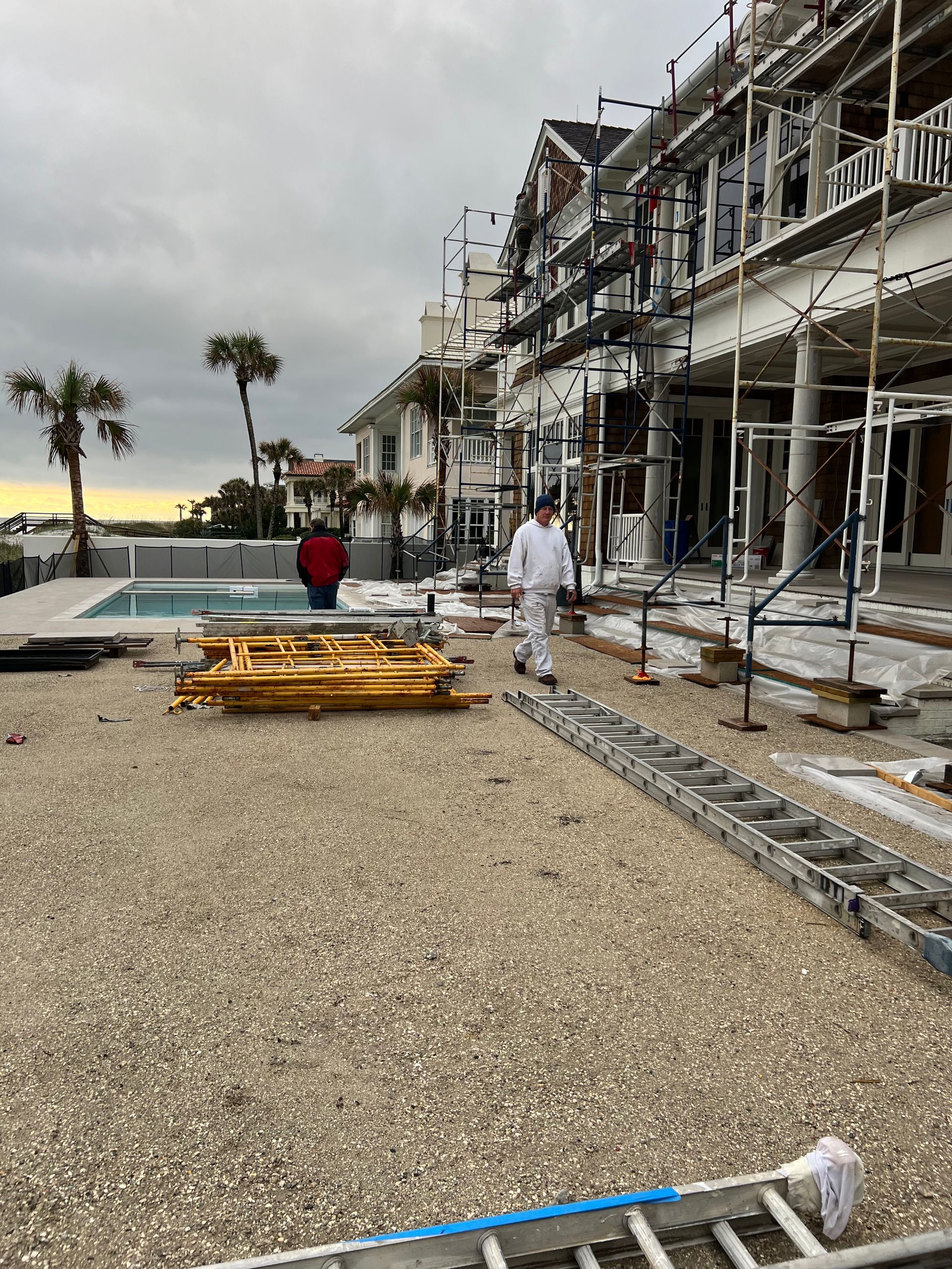 Construction site with a large house, scaffolding, workers, pool, and gravel. Cloudy day.