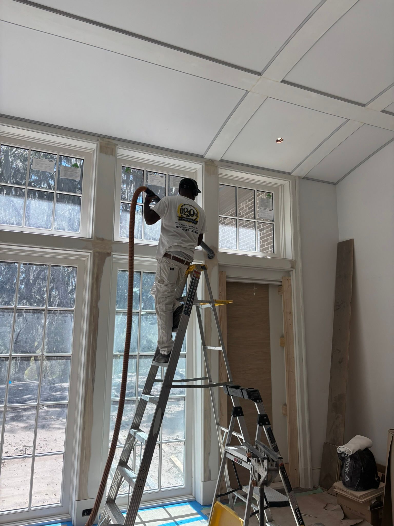 Worker on a ladder painting a tall window inside a bright room with white walls and ceiling tiles