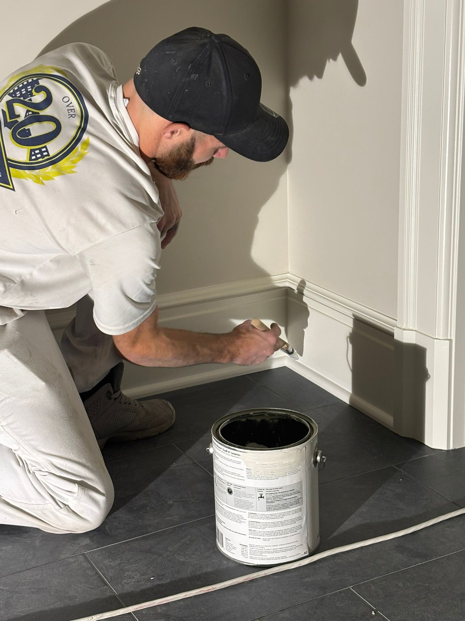 Worker painting white trim along a wall with a paint can on the floor