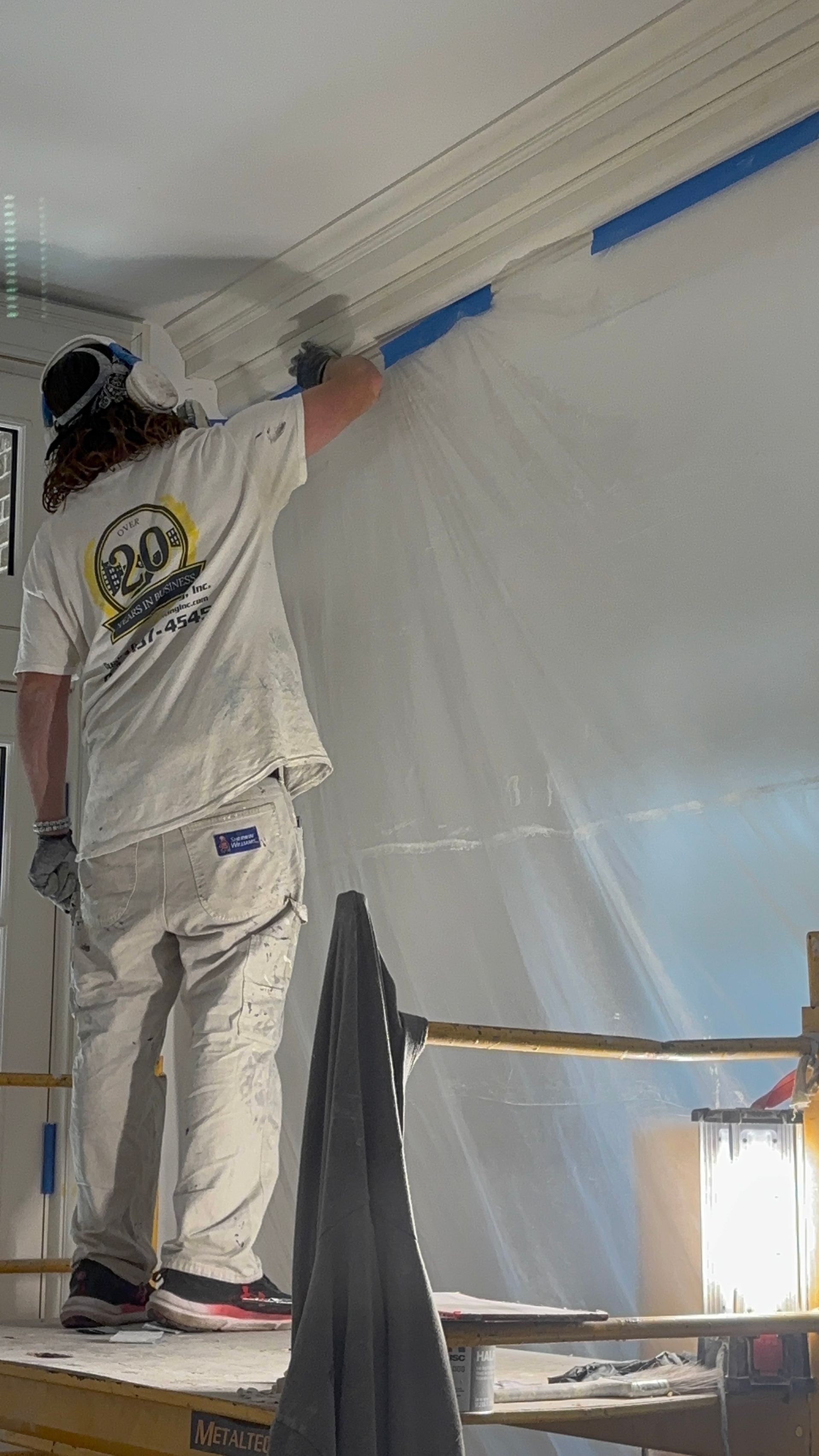 Painter on a ladder applying blue painter’s tape along a wall near the ceiling