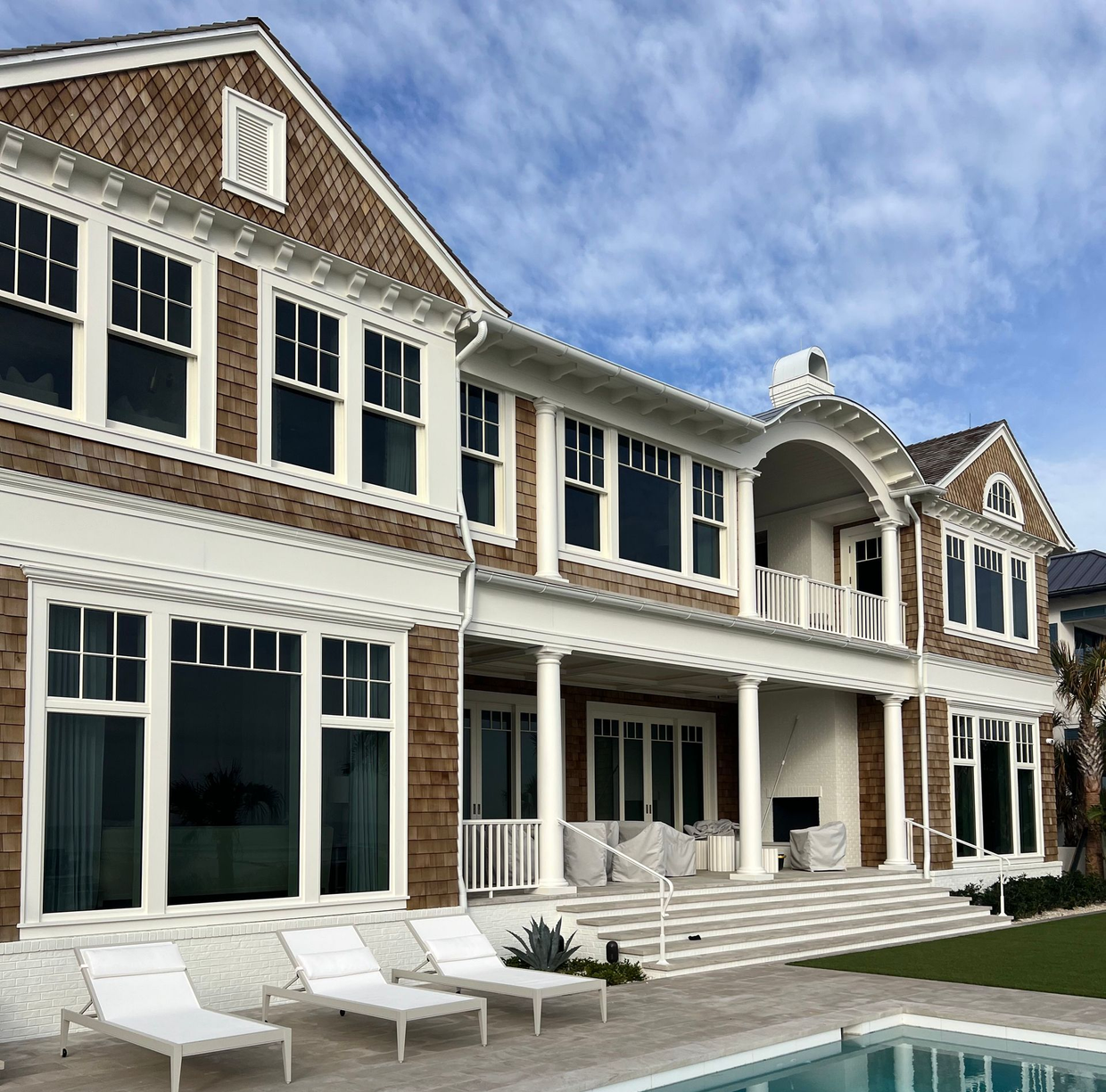 Luxury beachfront house with white lounge chairs and a turquoise pool under a partly cloudy sky