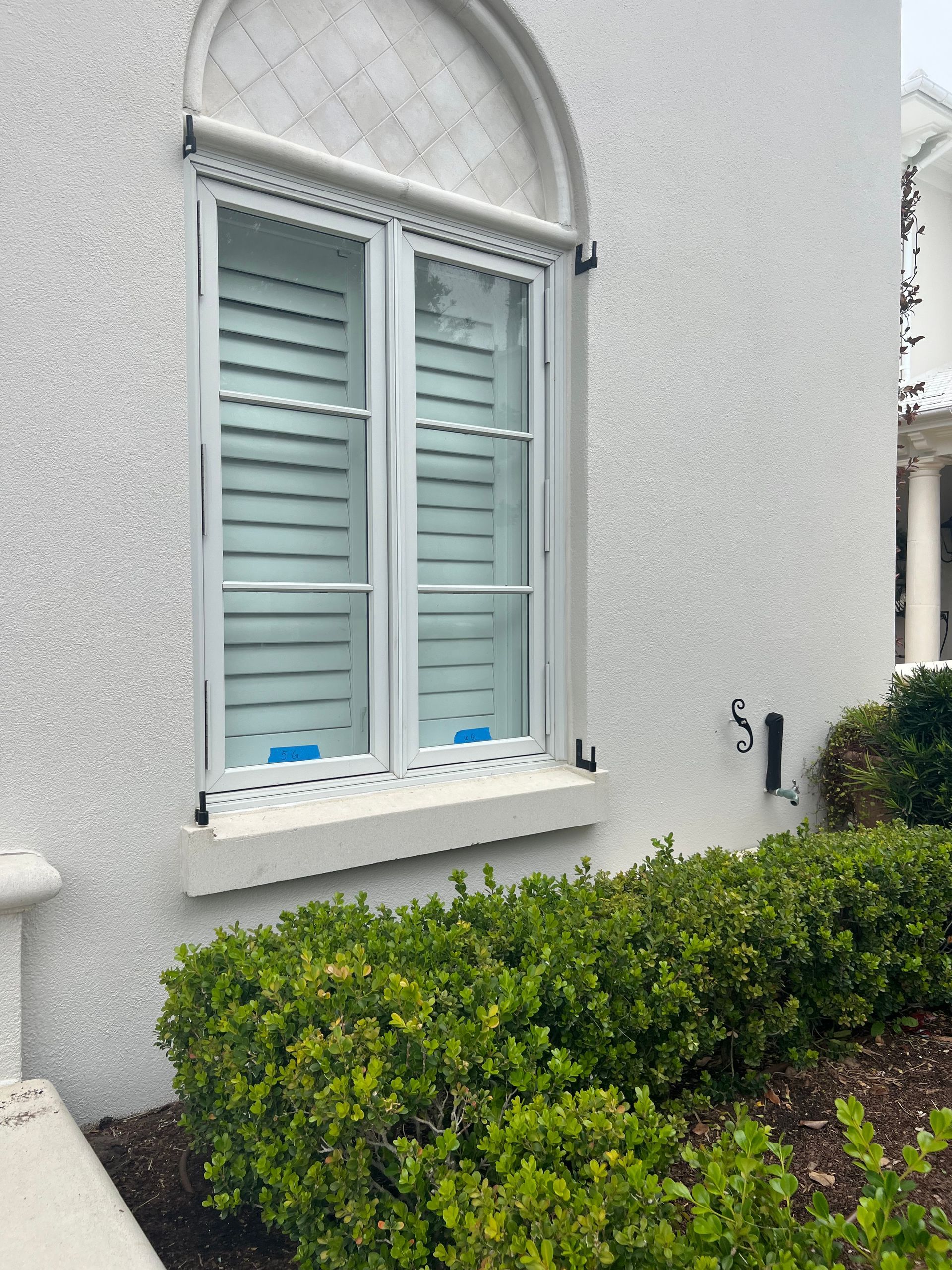 White window with closed light blue shutters, surrounded by white stucco and green shrubs.