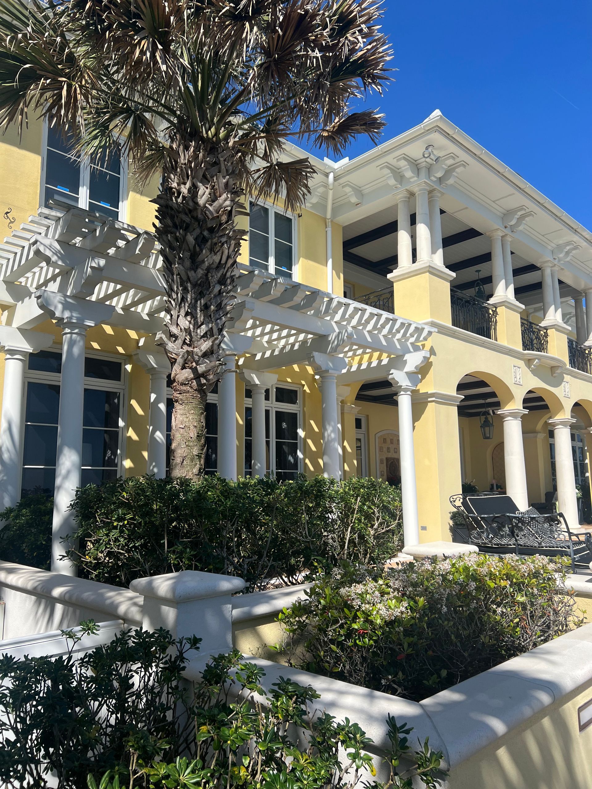 Yellow and white multi-story building with columns and arches, fronted by a low wall and shrubbery.