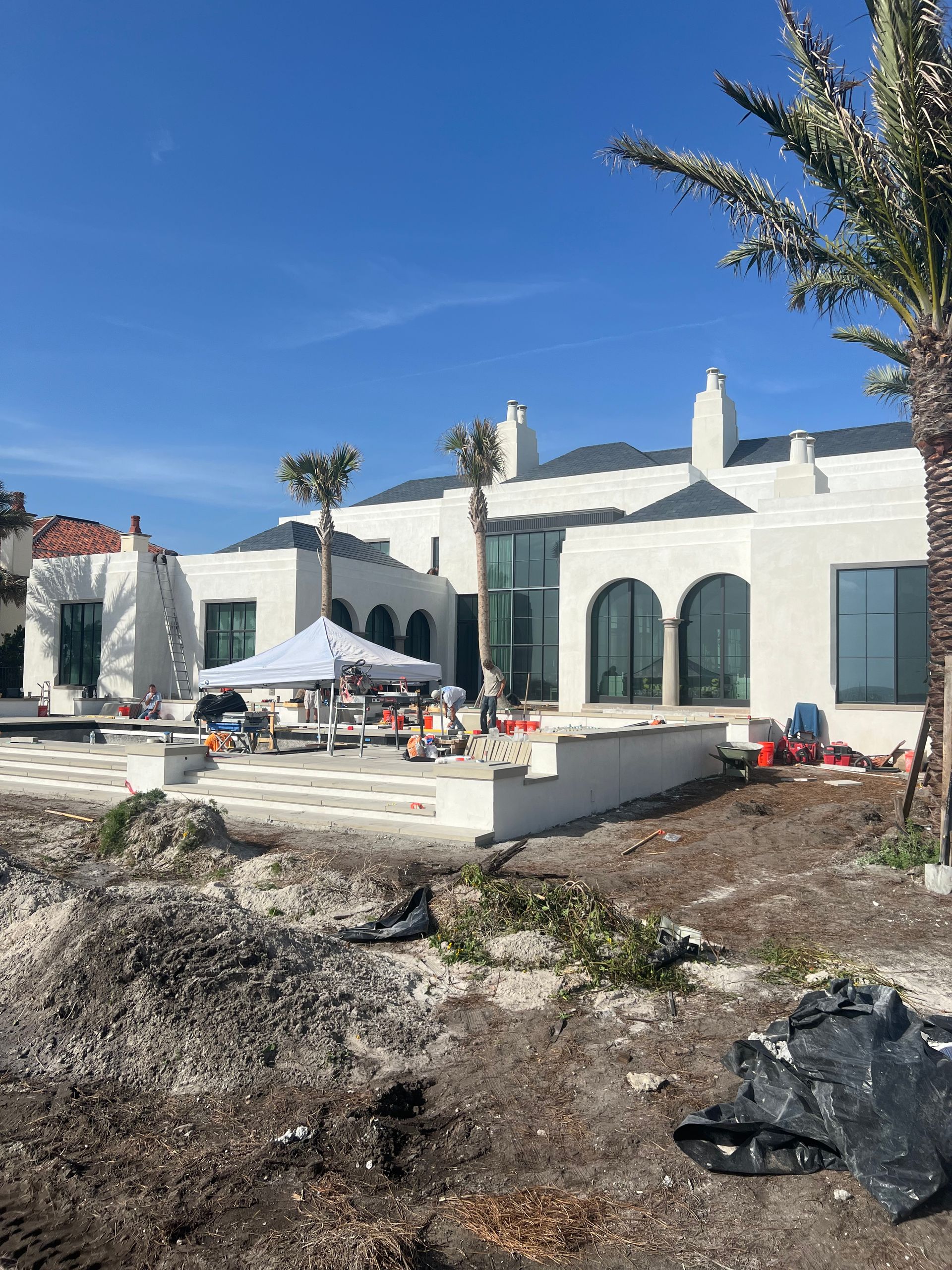 Exterior of a house under construction; white stucco walls, dark roof, workers, blue sky, and palm trees.
