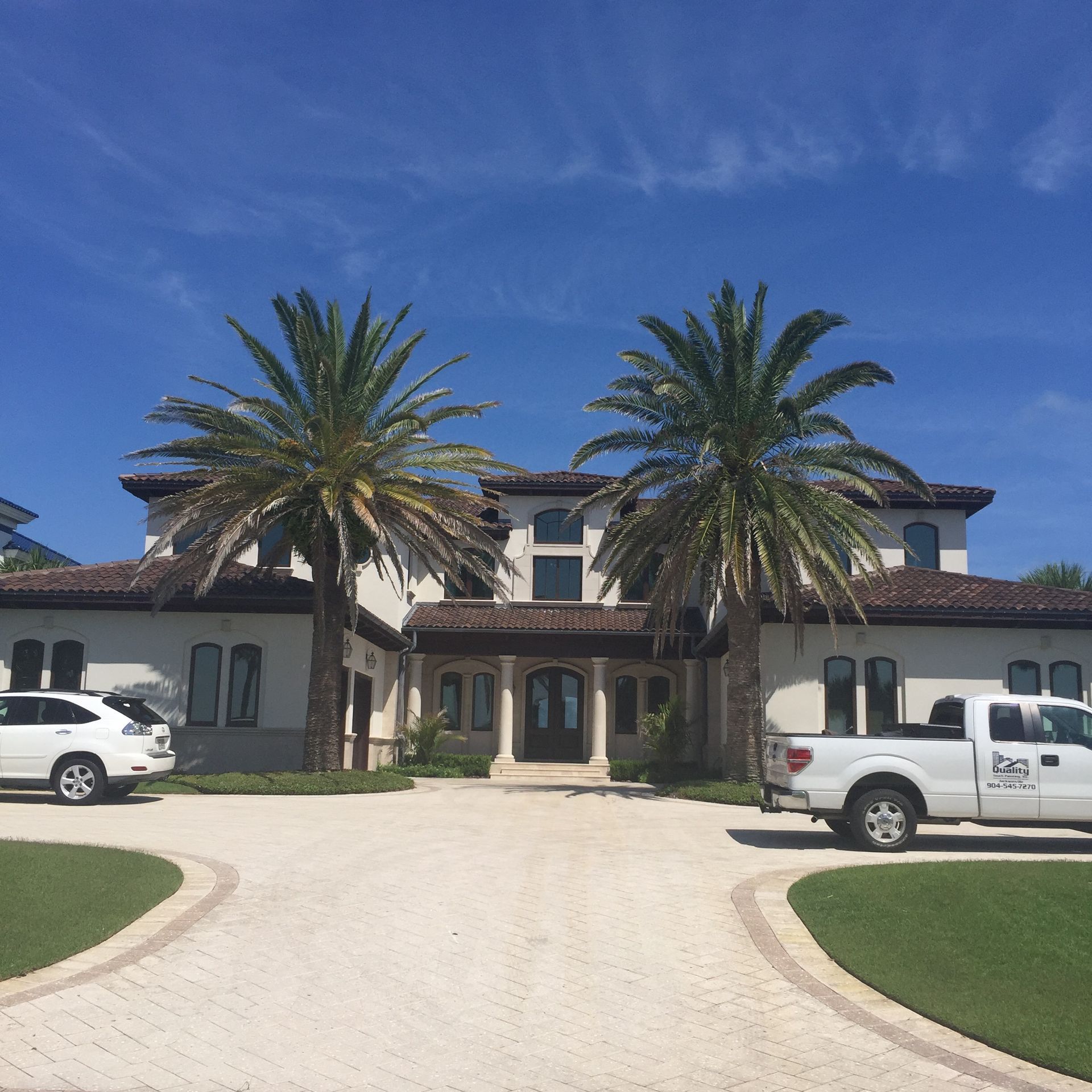 Large white house with a brick driveway, flanked by palm trees. Two white vehicles are parked in front. Blue sky.
