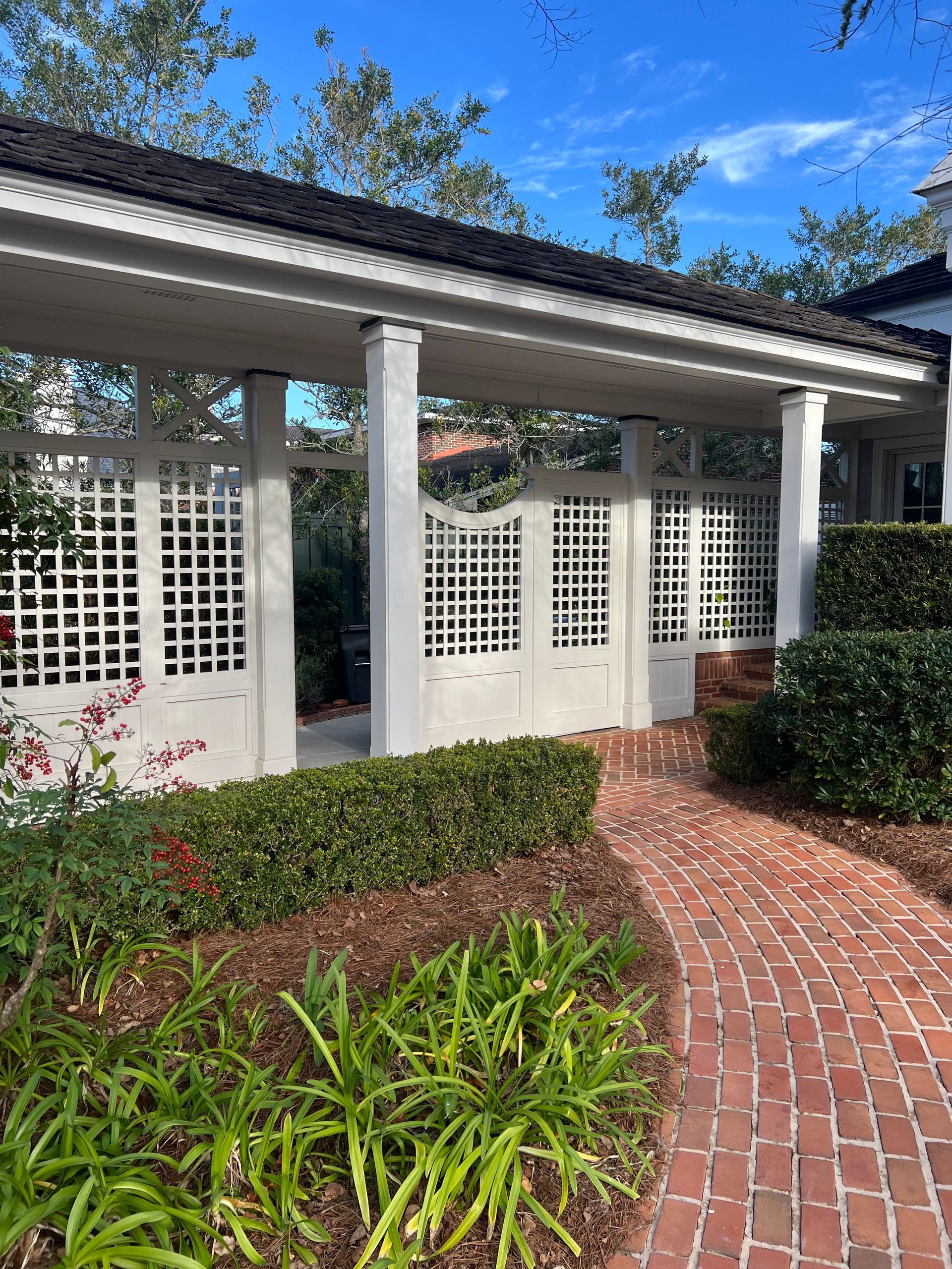 A brick pathway leads to a white lattice covered walkway. Green plants border the path and structure.