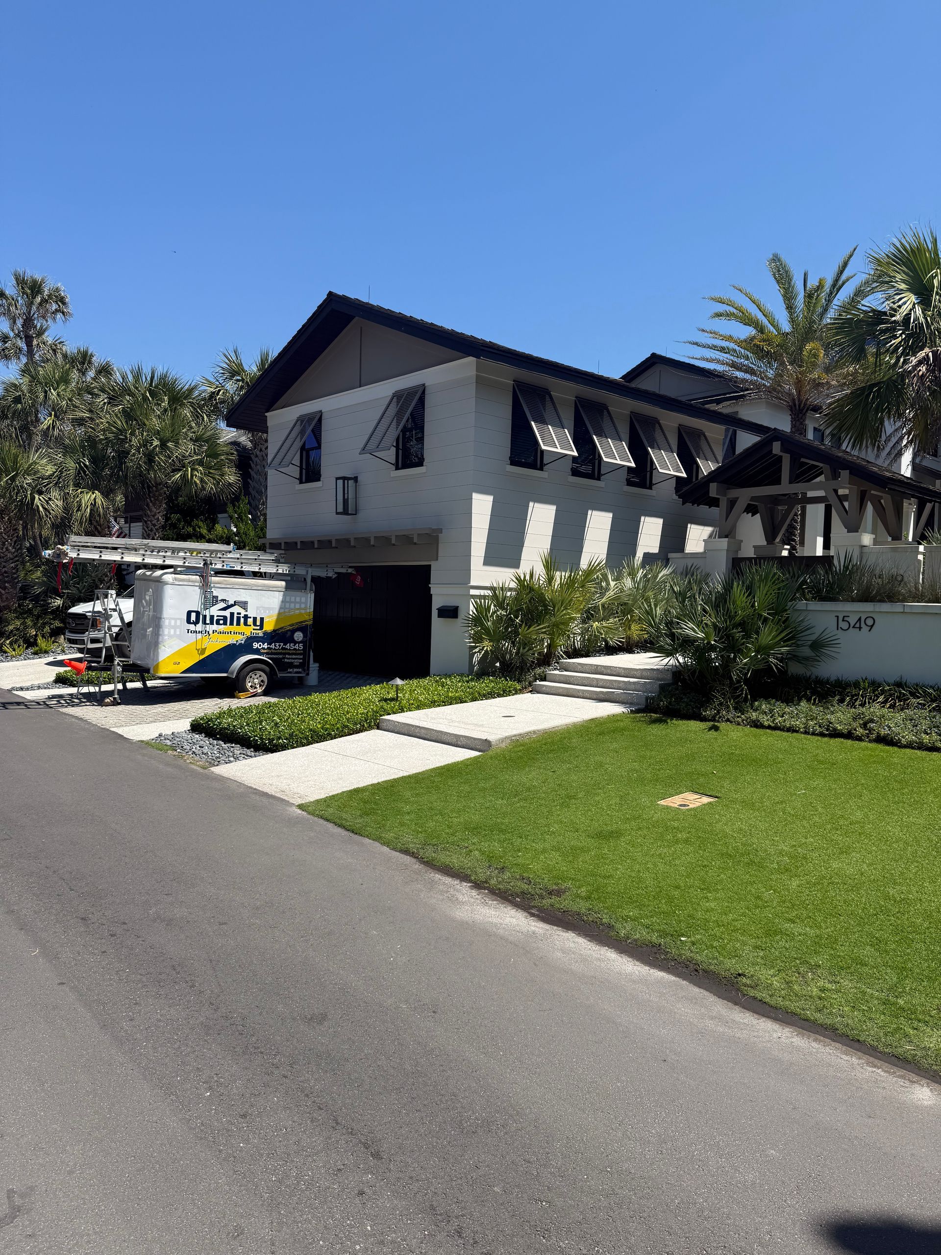 Two-story white house with black awnings, driveway, and moving van parked outside on a sunny day.