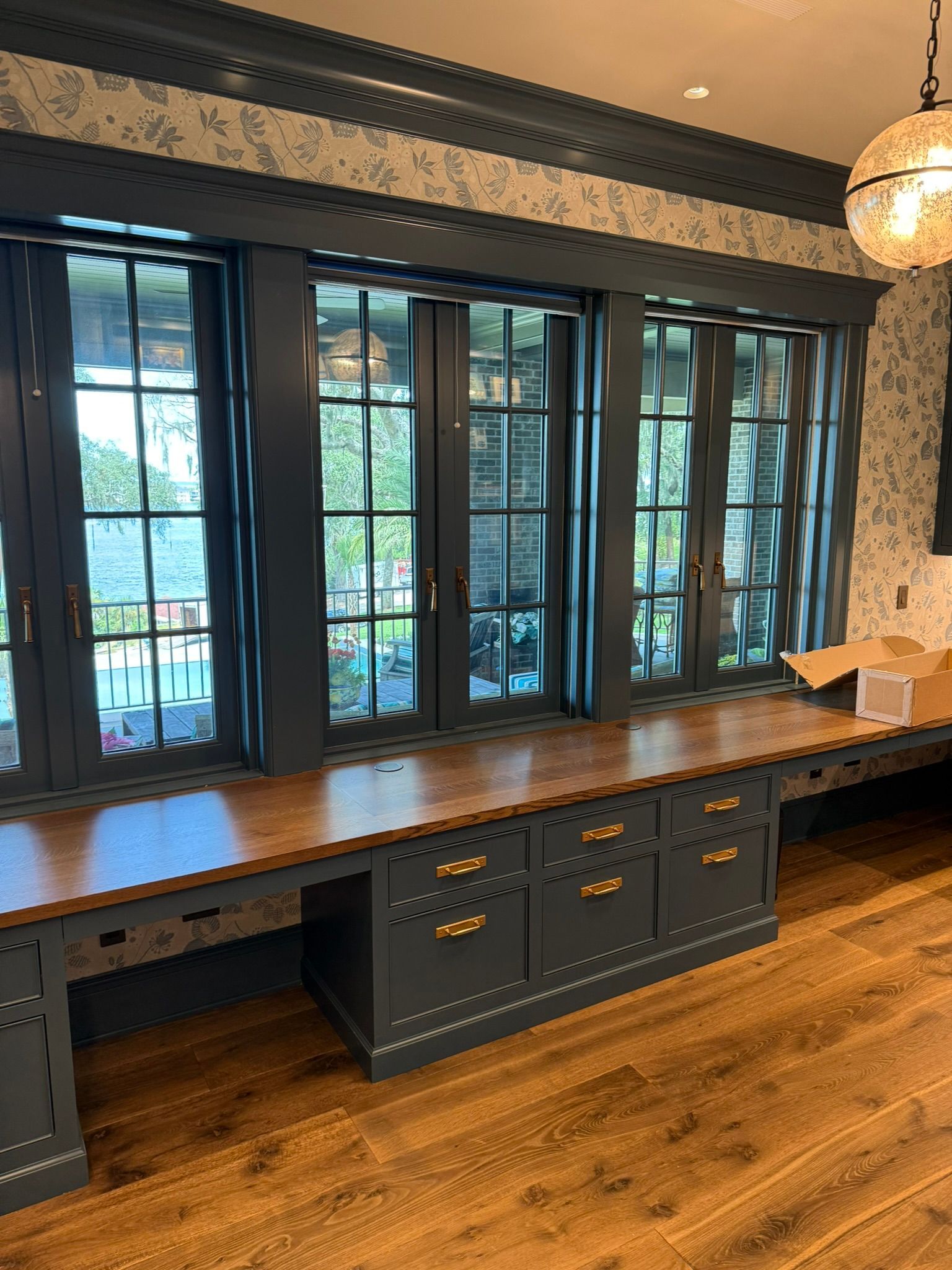 Built-in desk with dark blue cabinets, gold hardware, and wooden countertop below three windows.