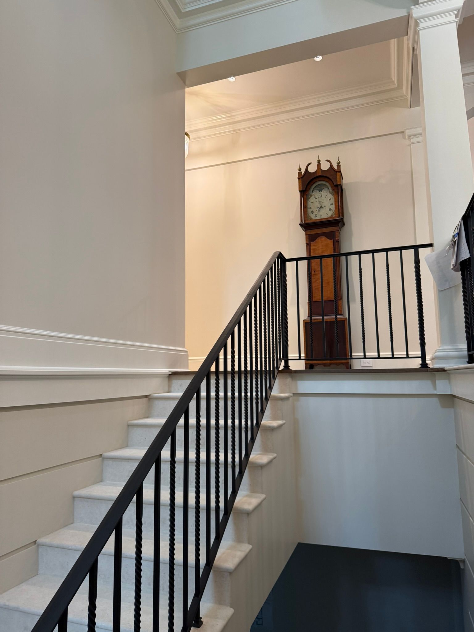 Indoor stairway with black railing leading to a landing and a tall grandfather clock against a cream wall