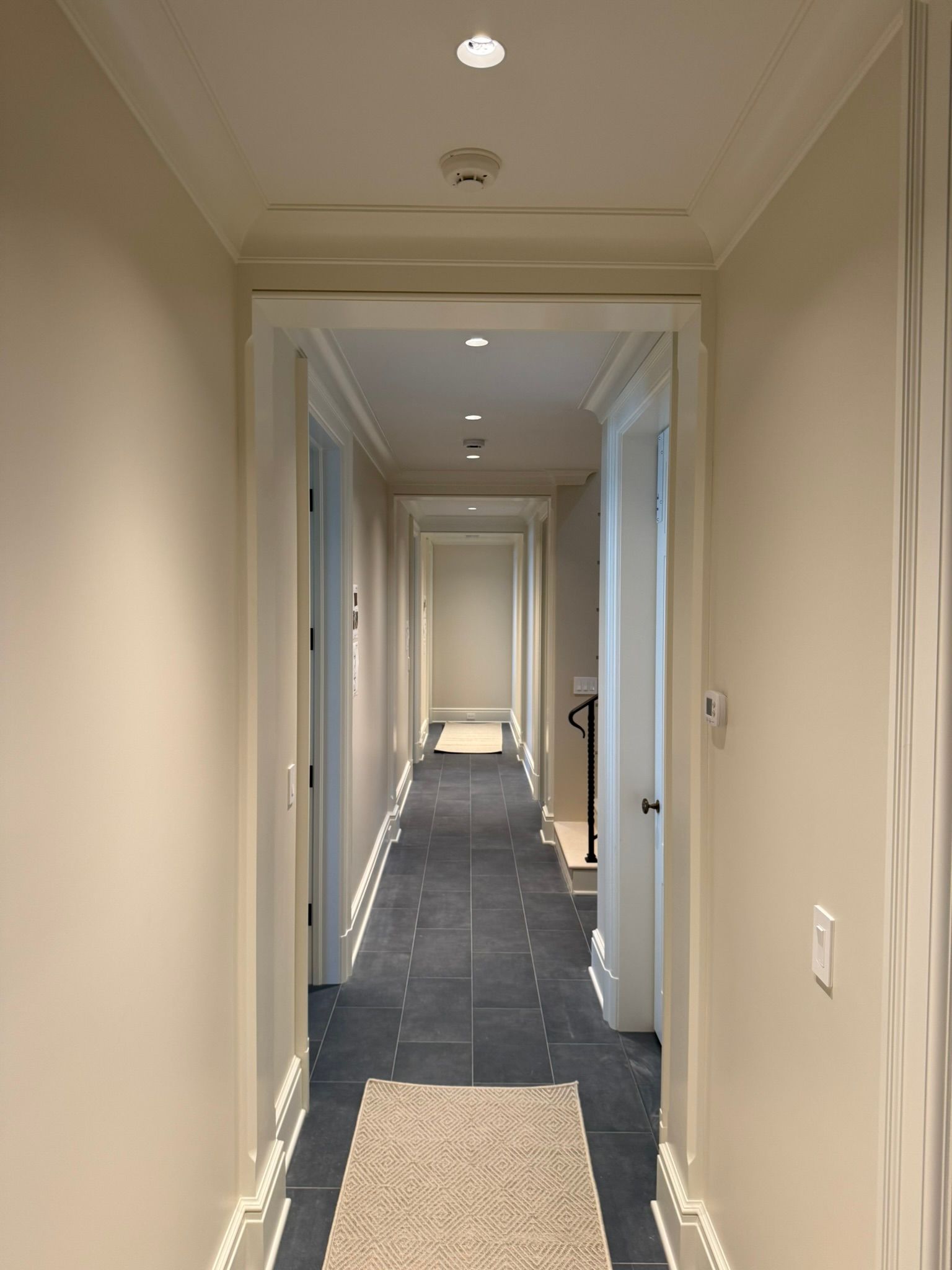 Long beige hallway with white doors, dark tile floor, recessed lights, and a runner rug.