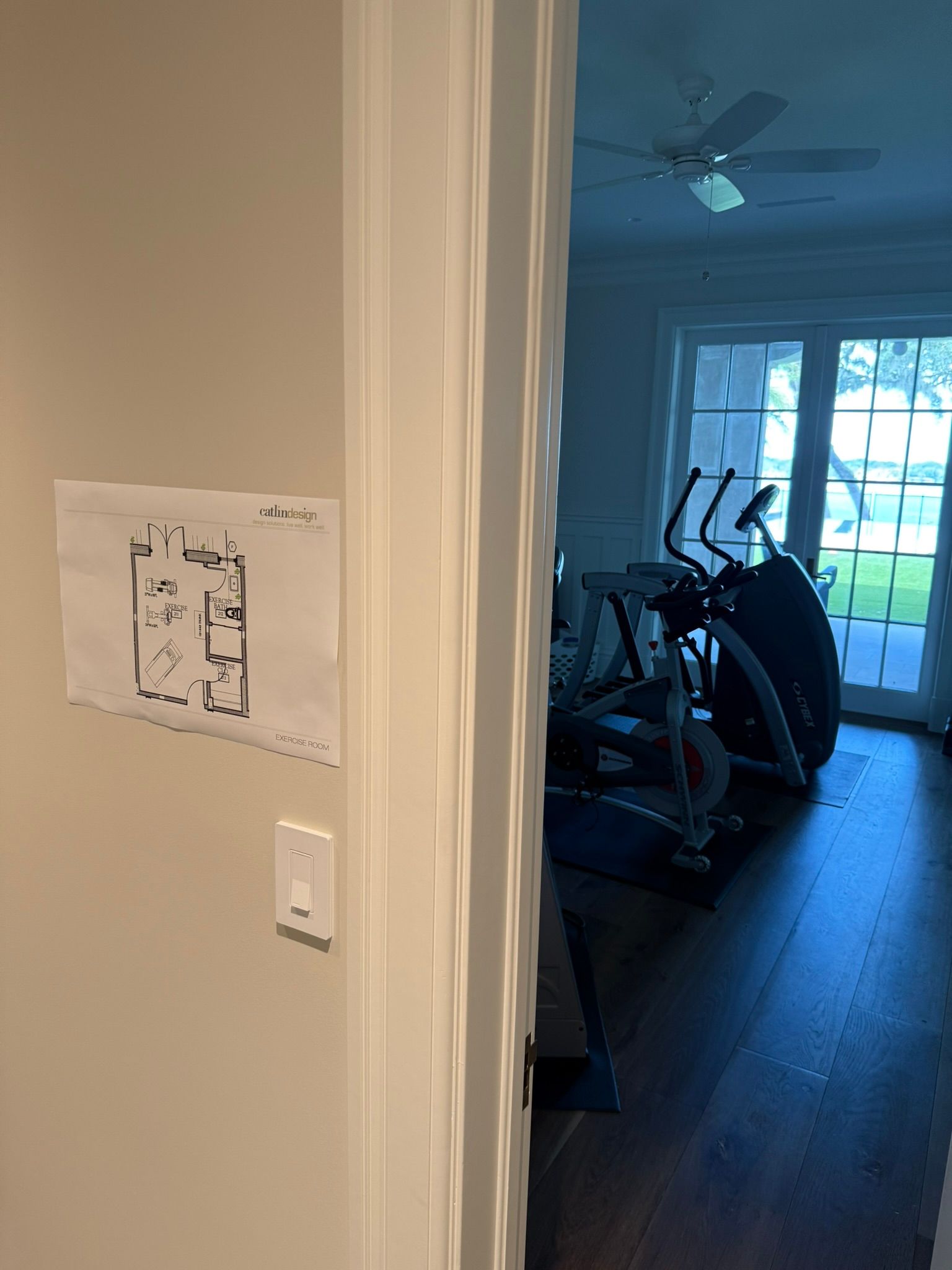 Hallway view into a room with exercise equipment and a glass door, with a paper taped to the wall in the foreground