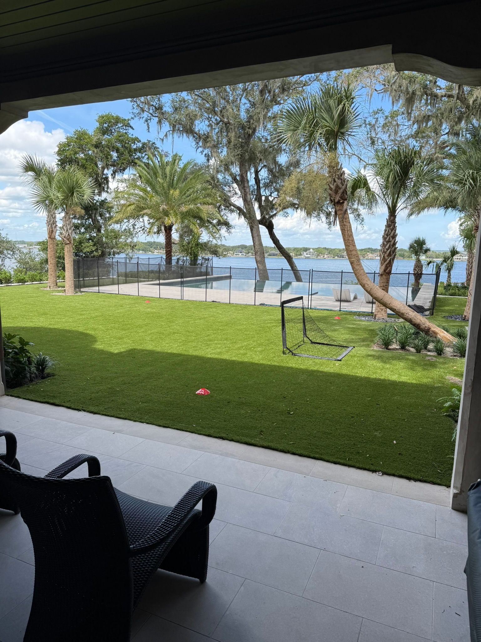 Porch view of a grassy yard with trees, a patio, and a calm waterfront under a bright sky