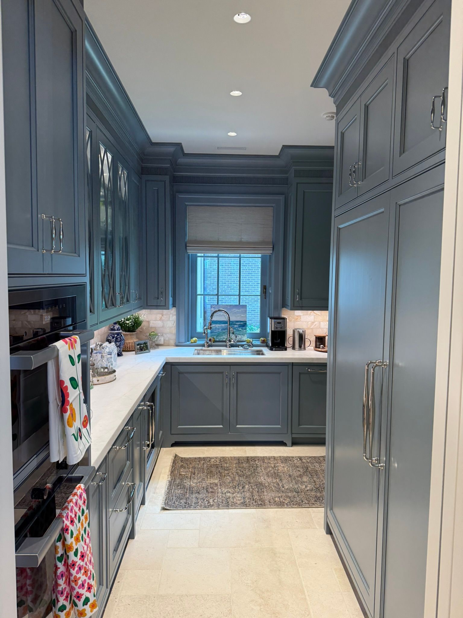 Narrow blue-gray kitchen with built-in cabinets, white countertops, window sink, and a patterned rug.