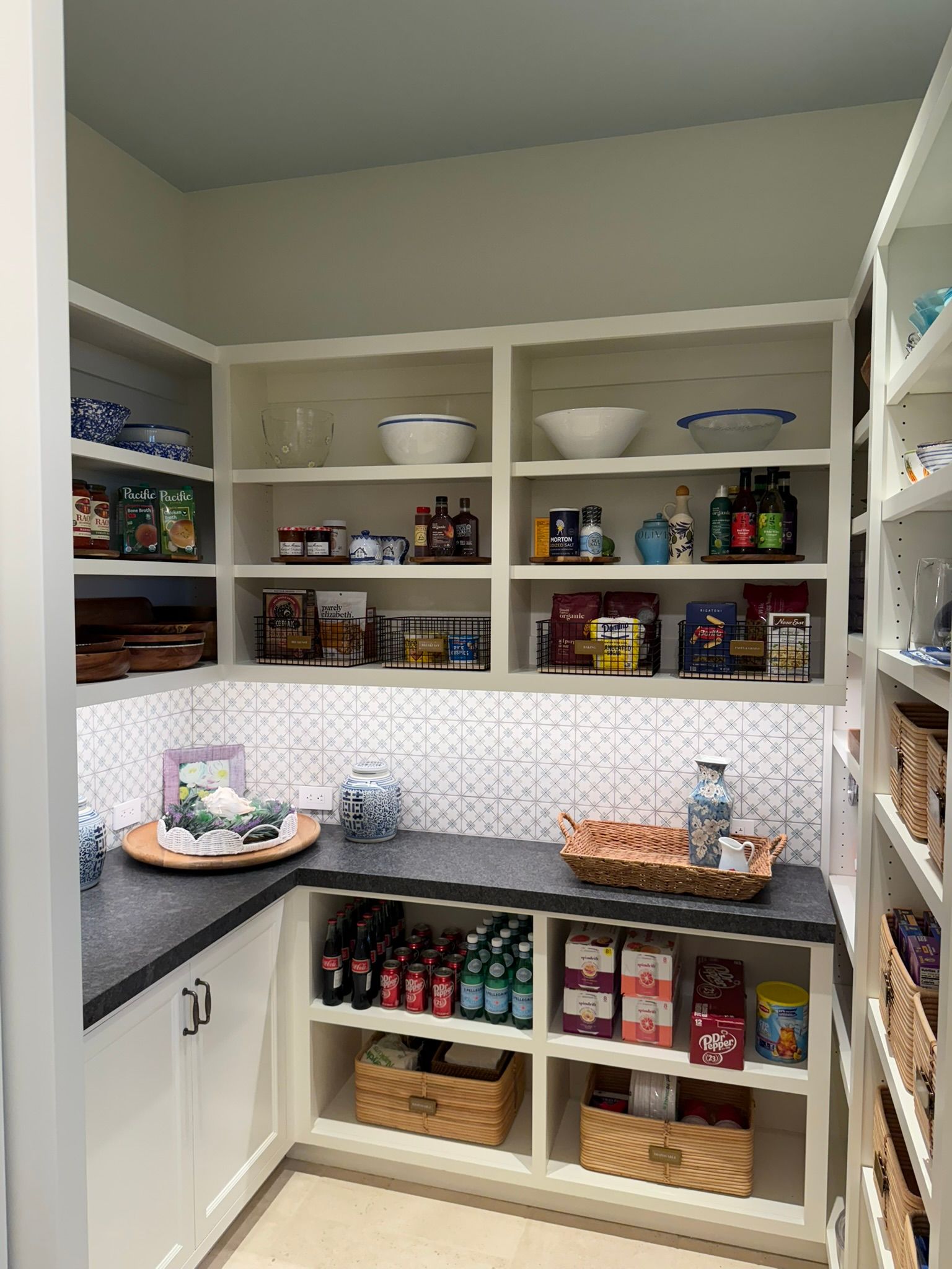 White pantry shelves with organized jars, boxes, baskets, and dishes in a corner kitchen.