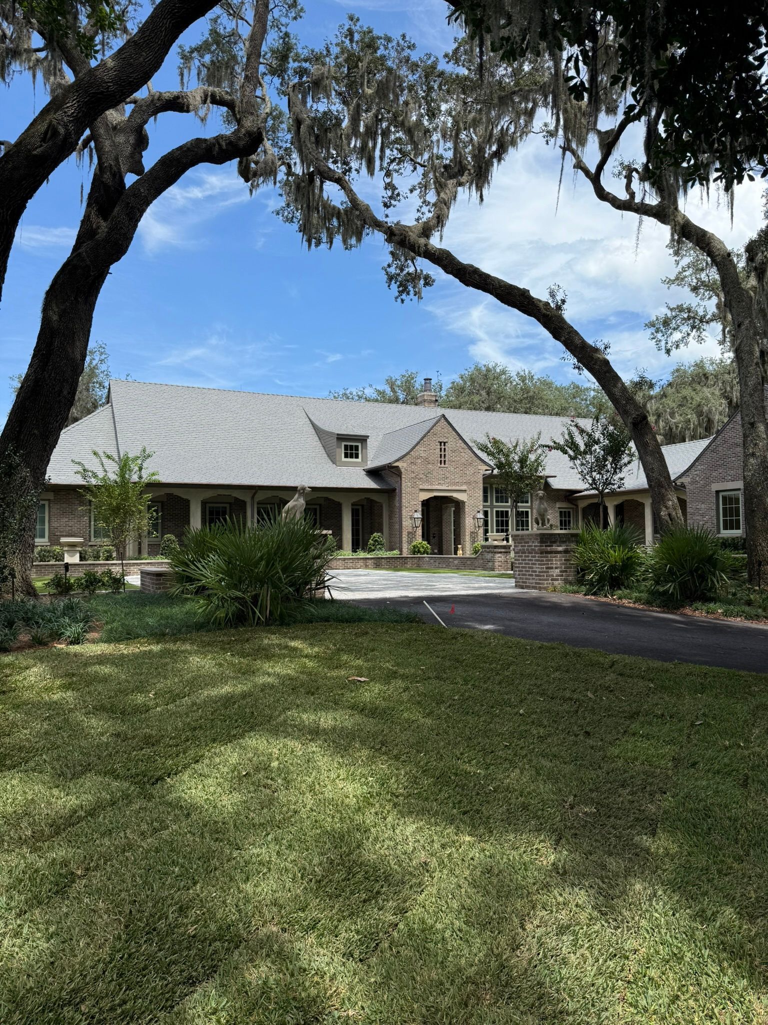 A one-story brick house with a stone facade, shaded by large trees, on a sunny day.