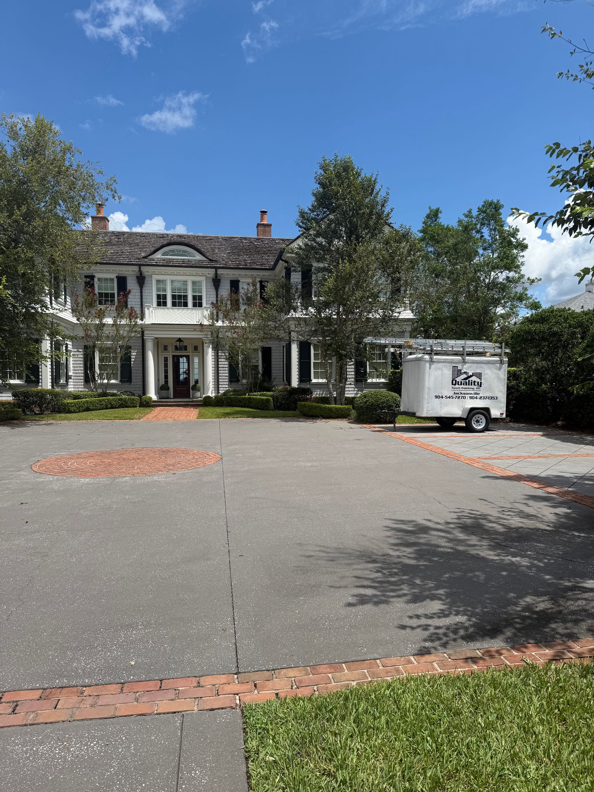 Large, two-story house with white trim, brick driveway, and service trailer parked nearby on a sunny day.