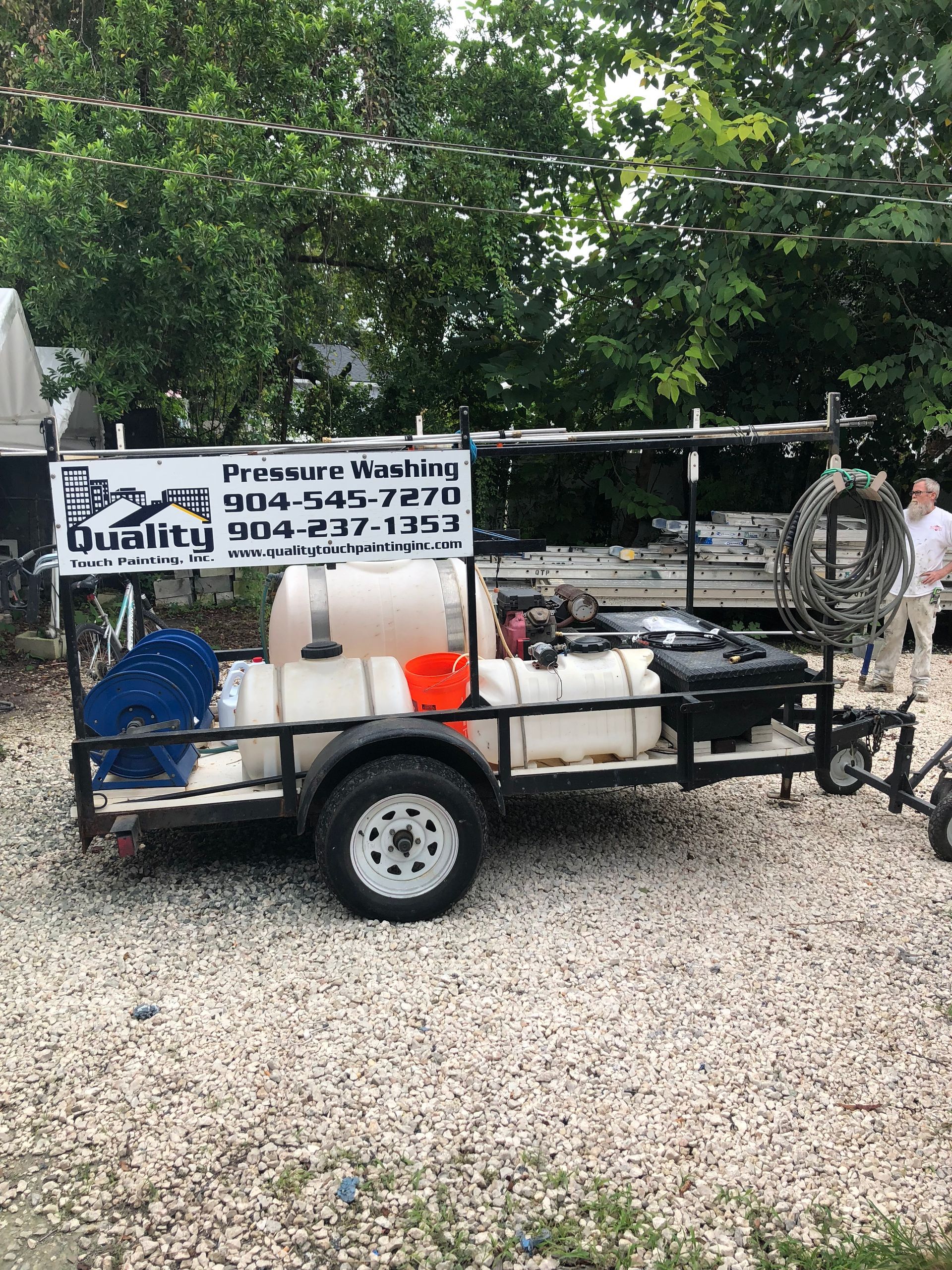 A trailer with a pressure washing setup; white tanks, blue hose reel, company sign, gravel.