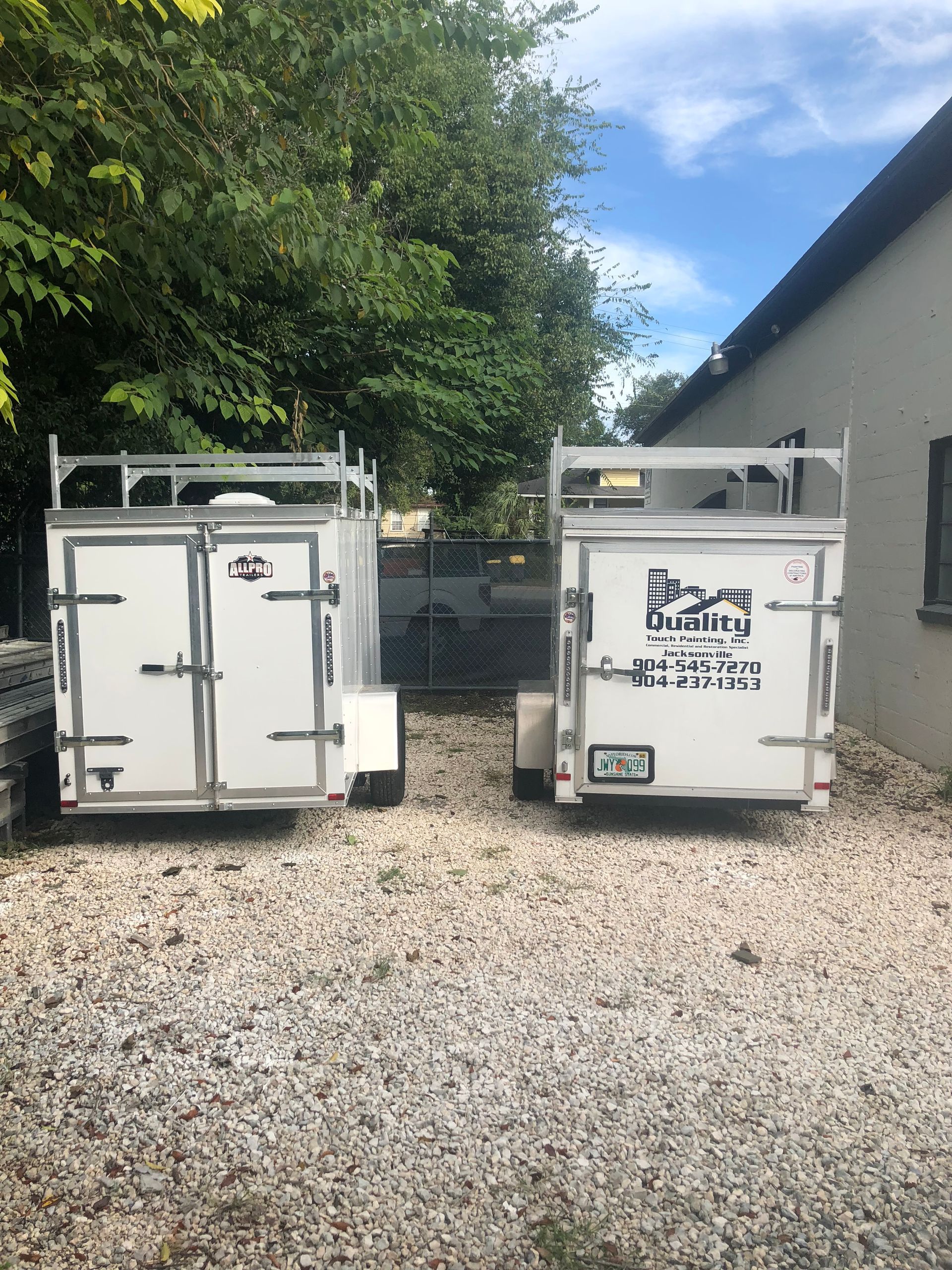 Two white enclosed trailers parked on gravel, with company logos, under a blue sky.