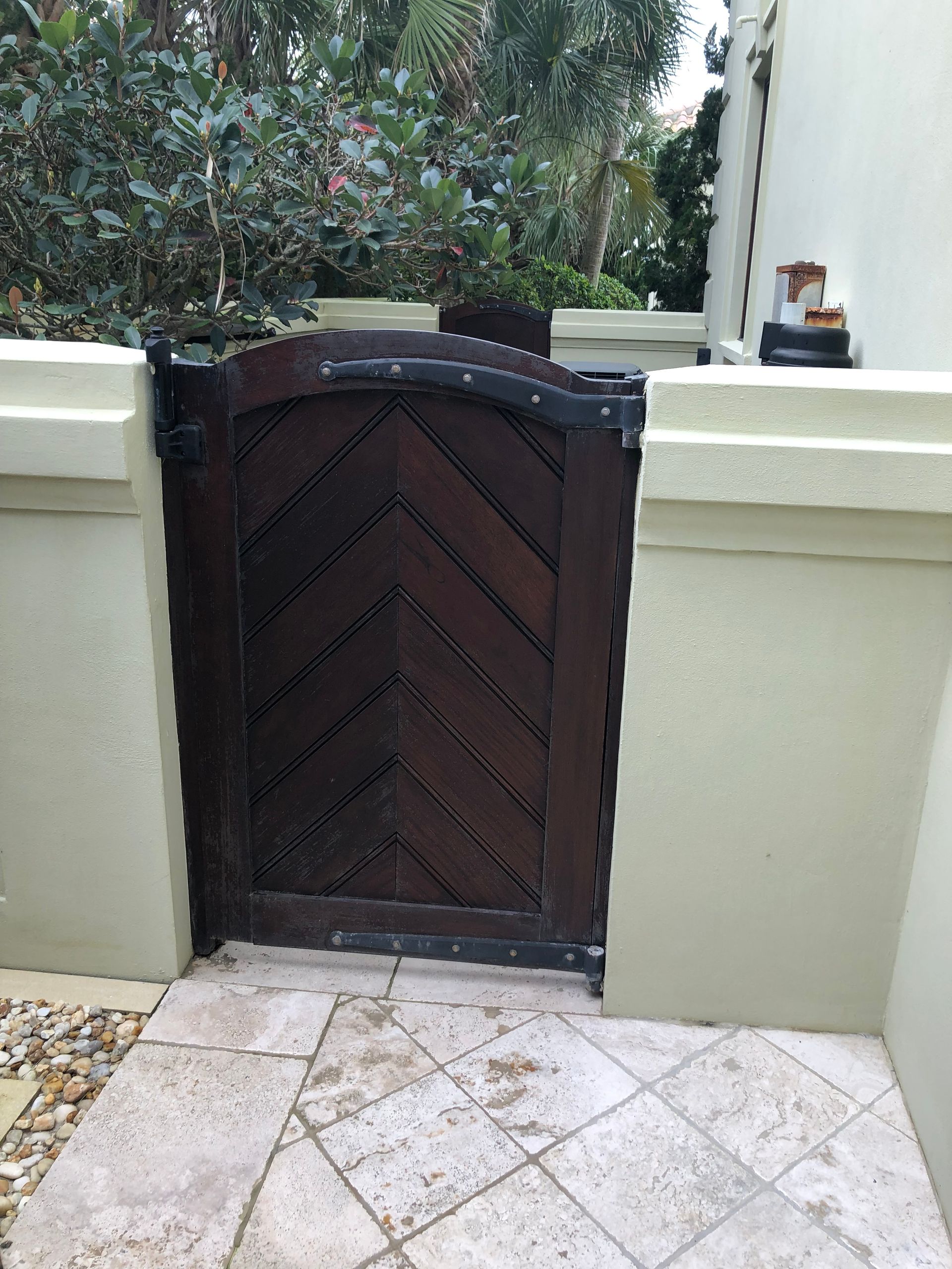 Wooden gate with chevron design, set in beige walls and paved walkway, with greenery in the background.