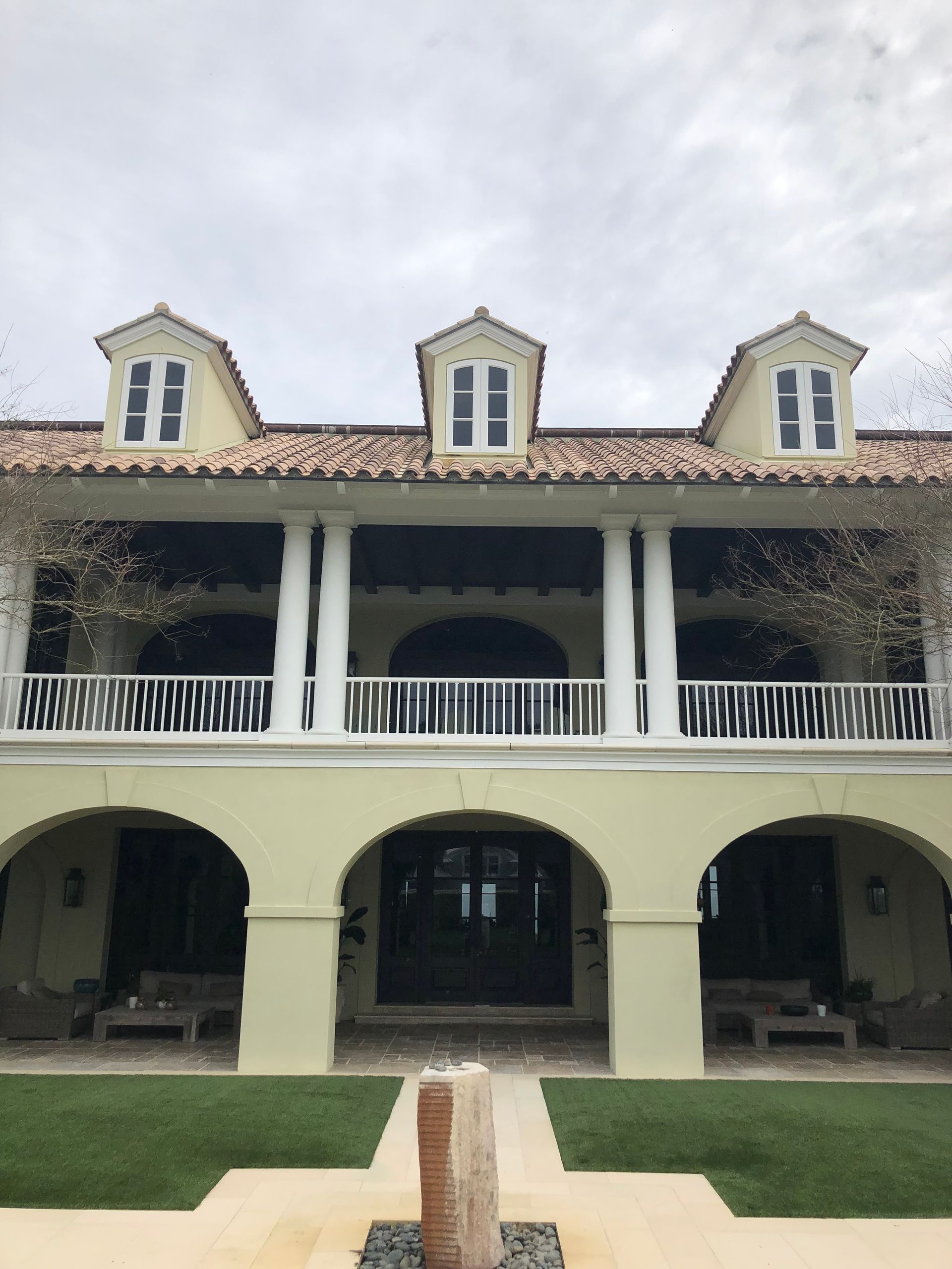 Two-story light yellow building with arches and columns, topped with three dormers, under a cloudy sky.