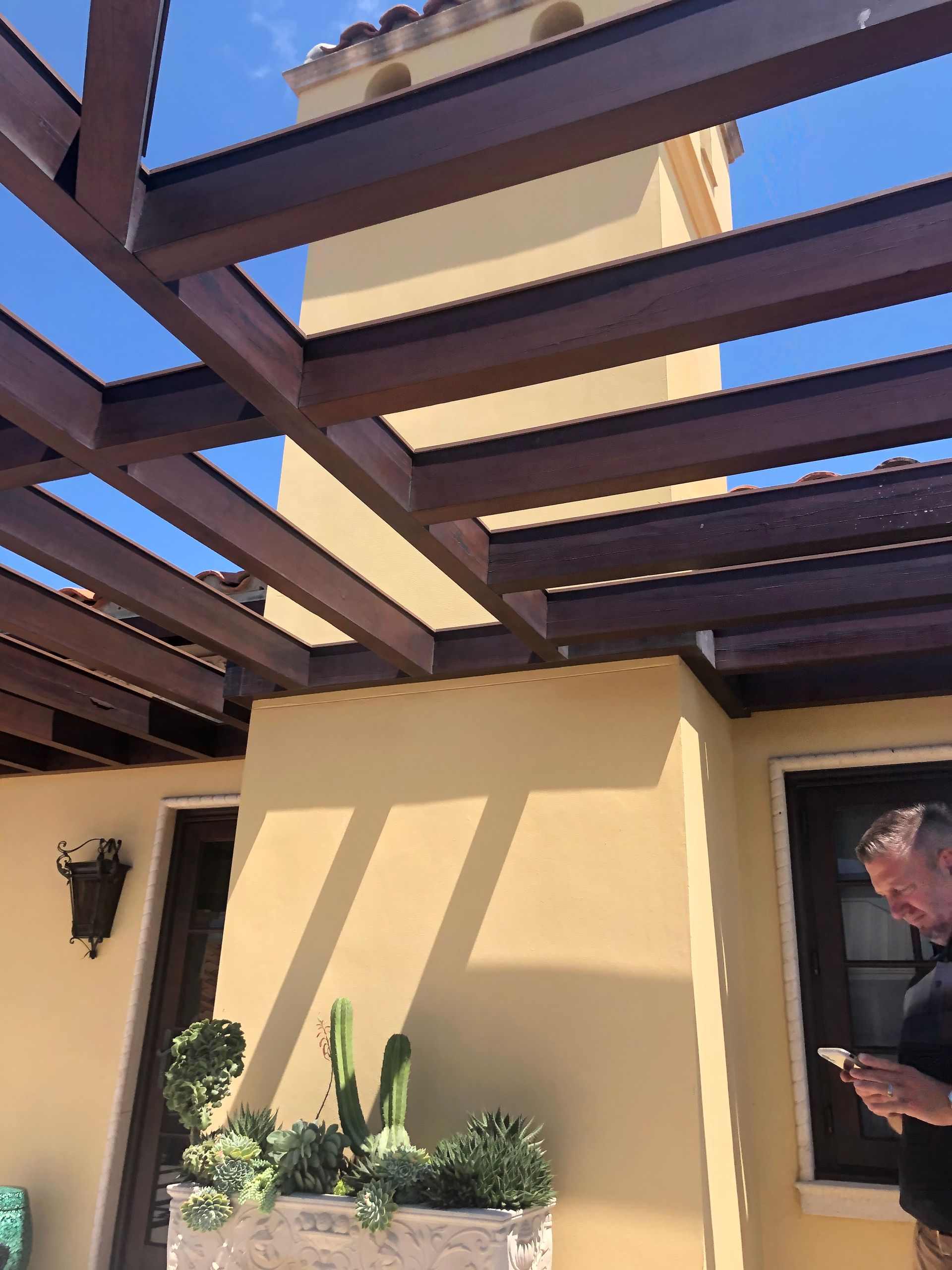 A pergola with dark beams casts shadows on a stucco wall, potted plants, and a man holding a phone.