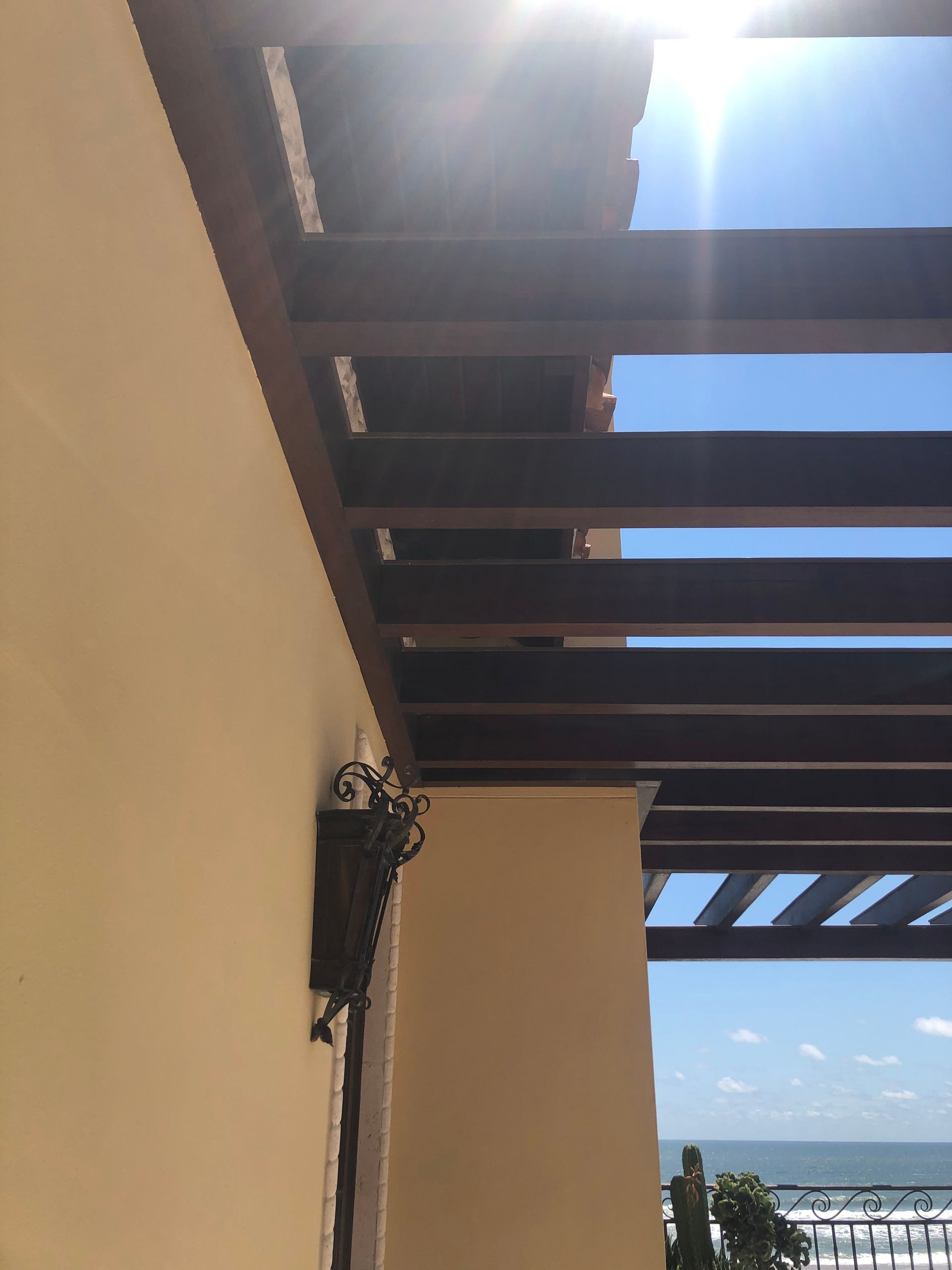 Pergola with brown wooden beams over a light yellow wall and ocean view. Bright sunlight.