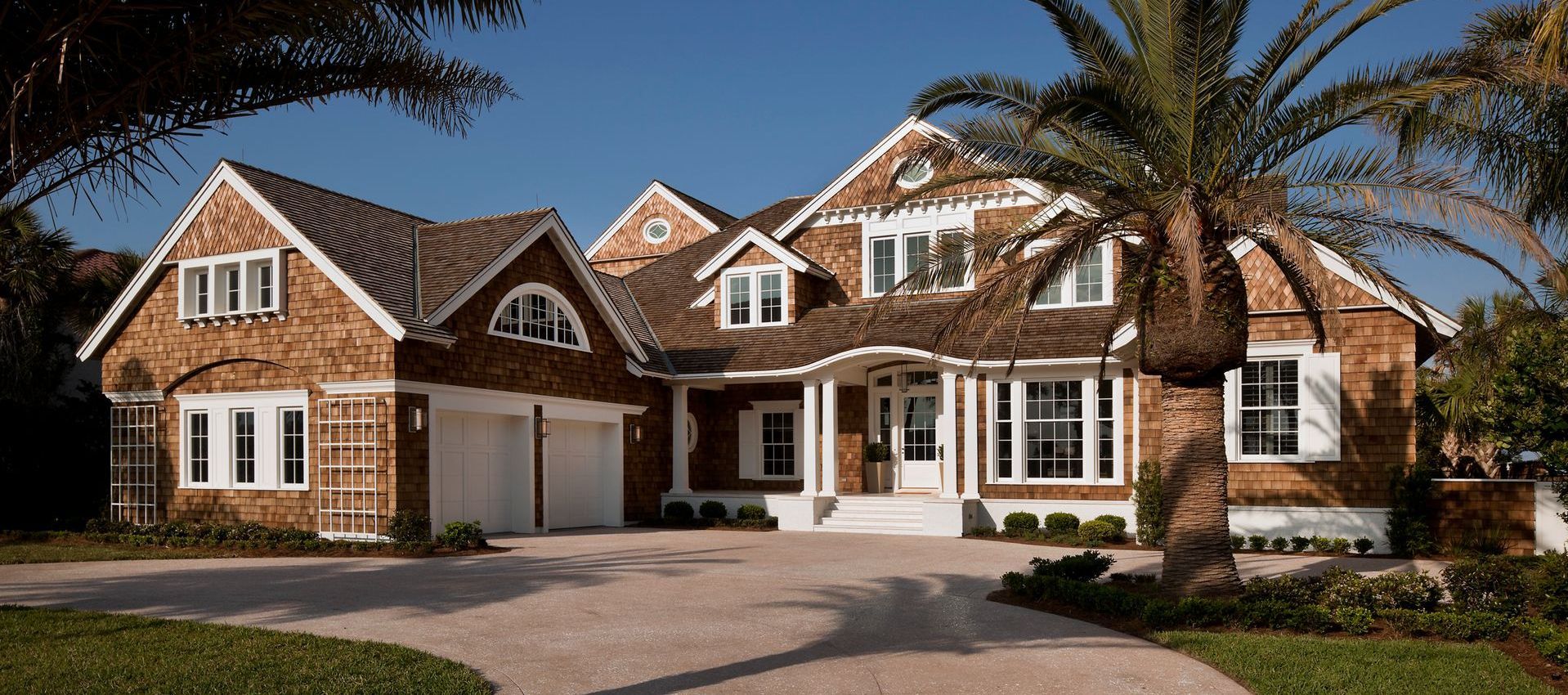 Large brick suburban house with white trim, multiple gables, and a two-car garage under palm trees