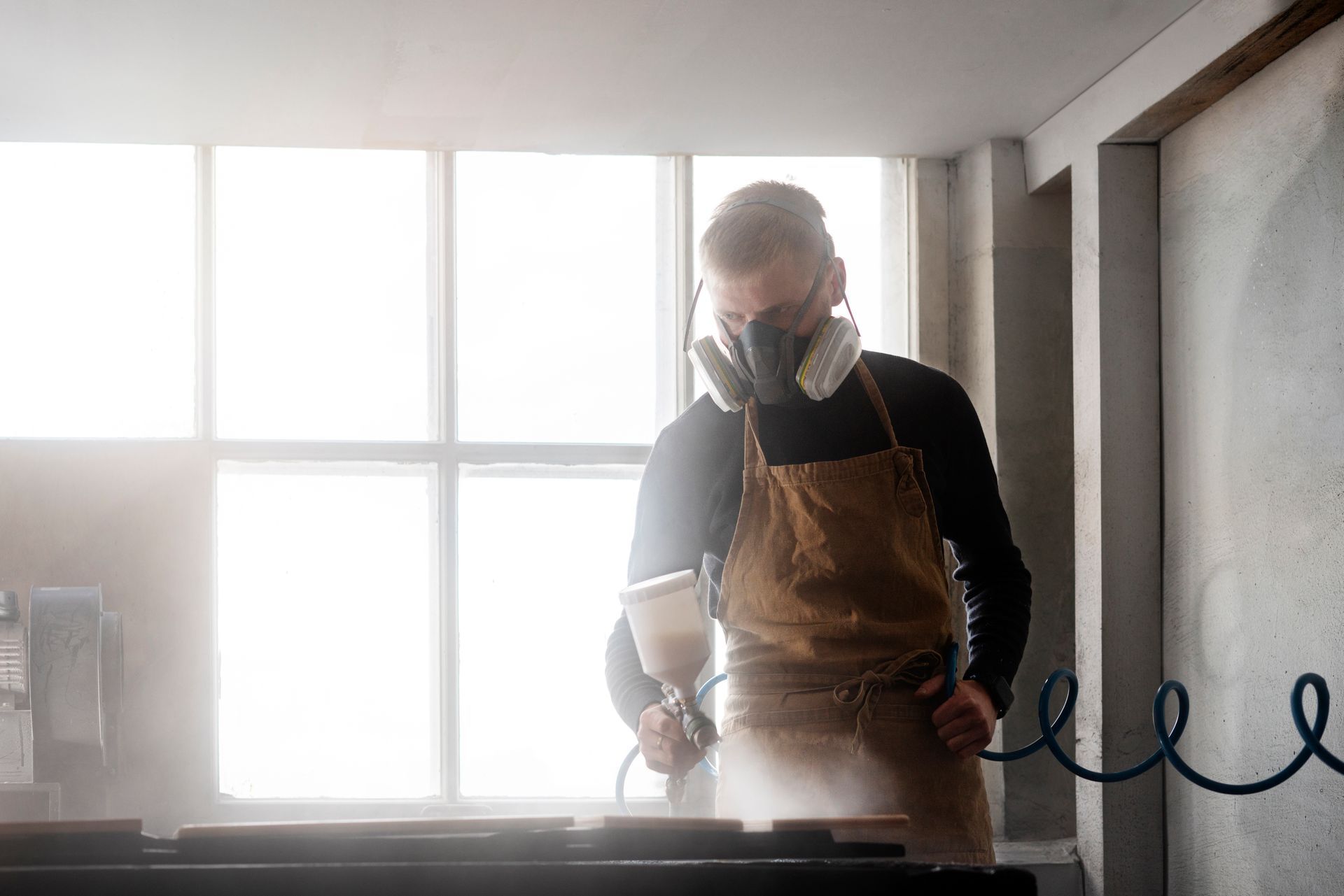 Person wearing respirator mask sprays wood in workshop near window.