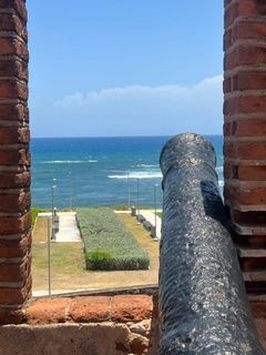 An antique black cannon points toward a coastal path leading to the blue ocean, viewed through a red brick embrasure.