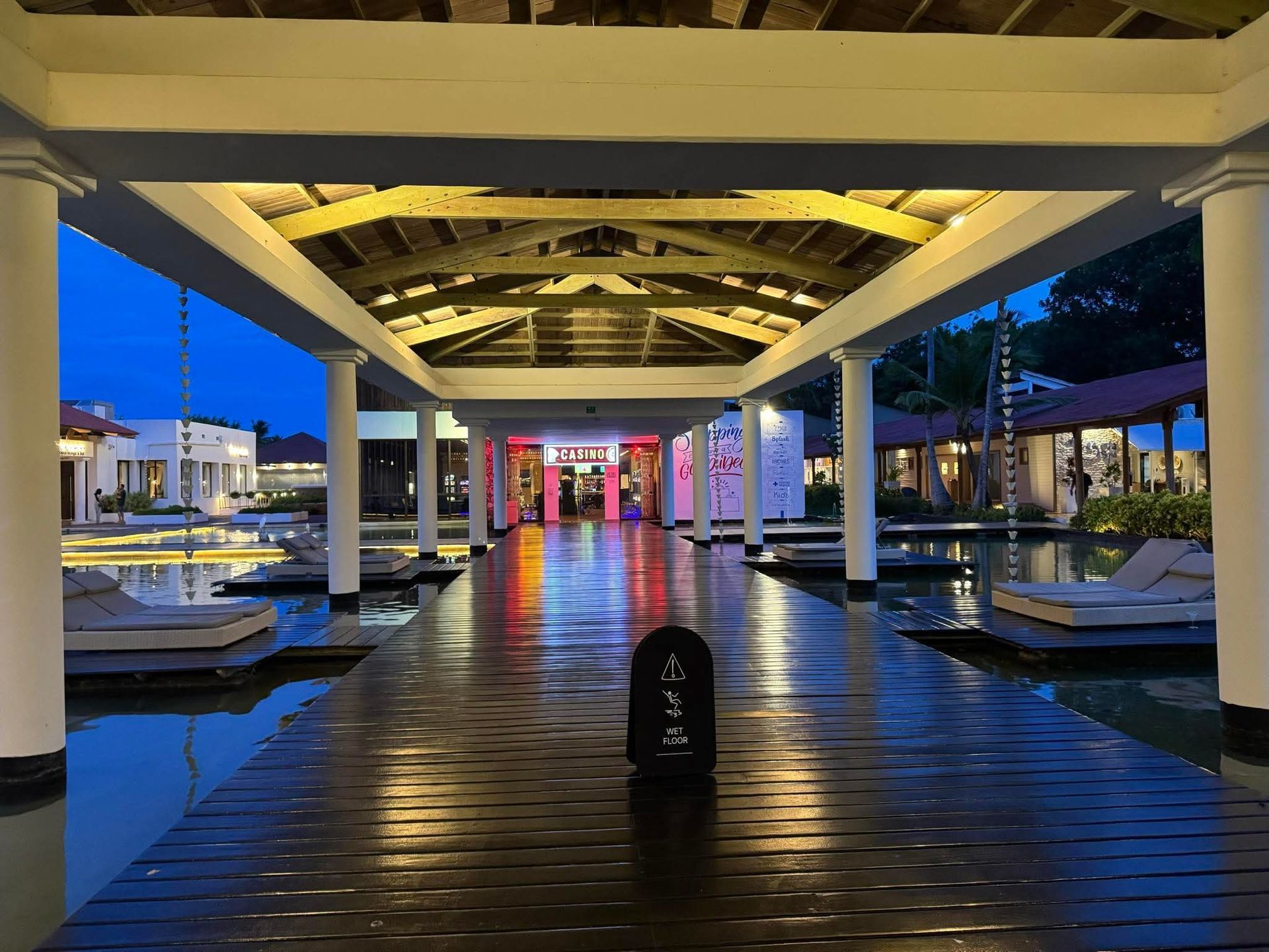 A wooden walkway over water leads through an illuminated, covered resort area at dusk, with lounge chairs on the sides.