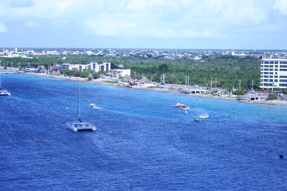 A coastal view of a blue sea with boats near a shoreline featuring buildings and trees under a cloudy sky.