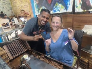 A person teaching another how to roll a cigar in a workshop with wooden tools and materials on a table.