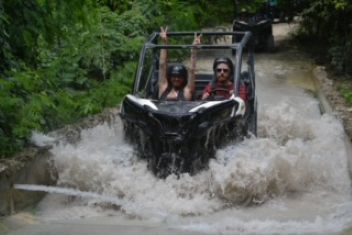 Two people in helmets ride a black off-road vehicle through deep water on a jungle trail, one signaling a peace sign.