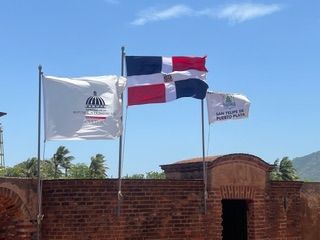 Three flags, including the Dominican Republic flag, fly above a historic red brick structure under a blue sky.
