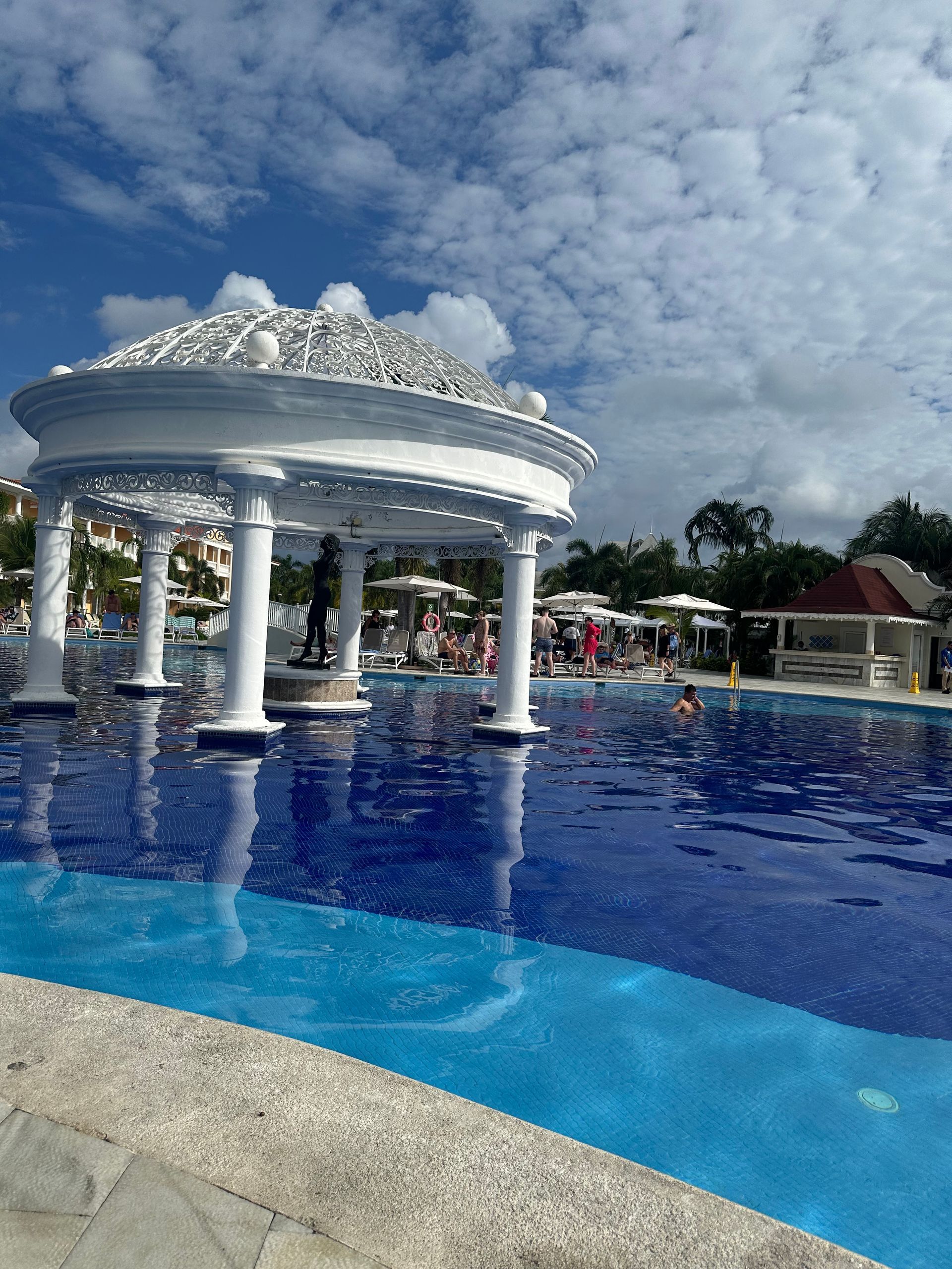 A white, ornate gazebo stands in the middle of a vibrant blue resort pool under a sunny, cloud-filled sky.