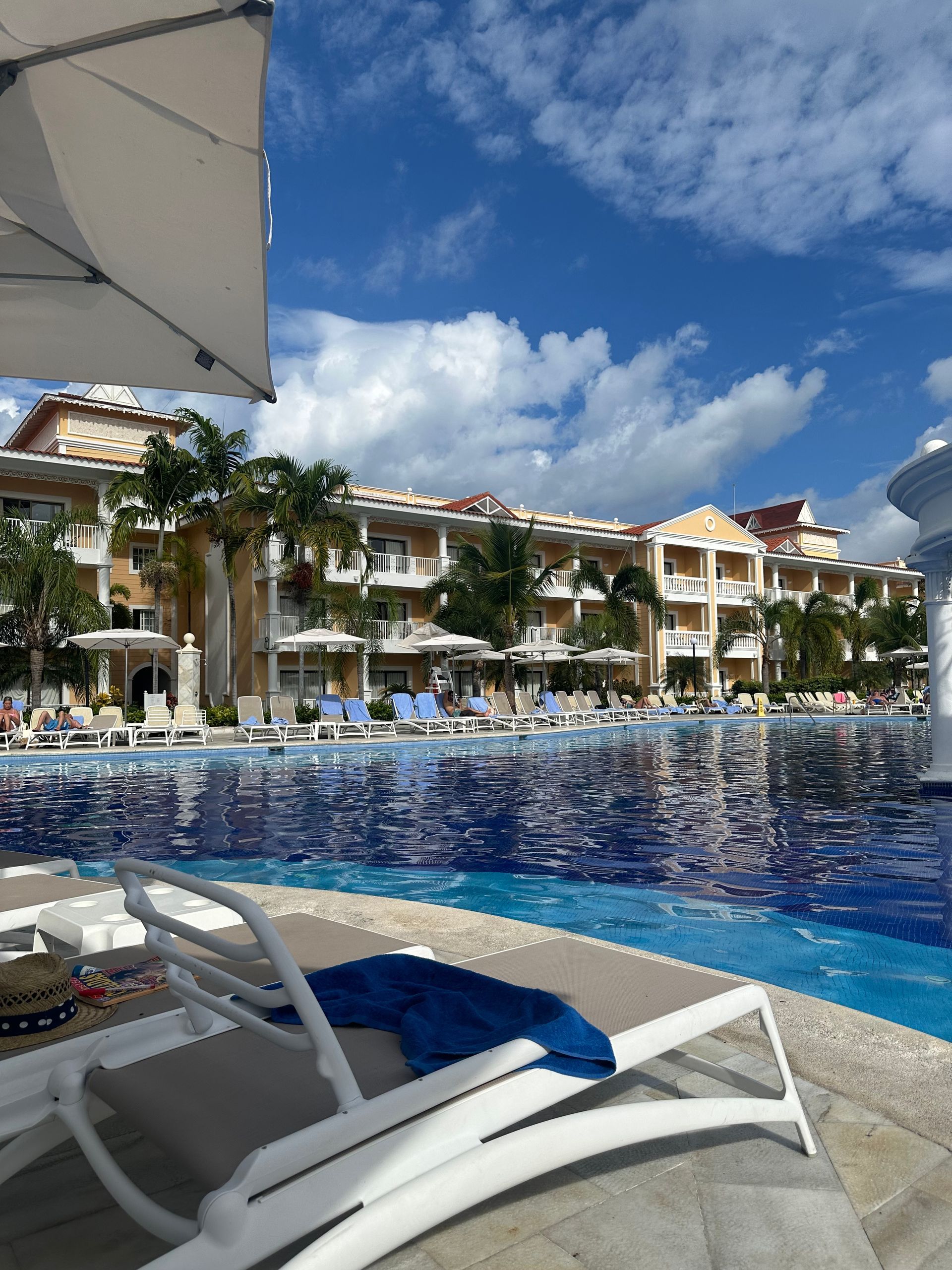 An empty lounge chair with a blue towel sits on a poolside patio facing a resort building under a bright blue sky.