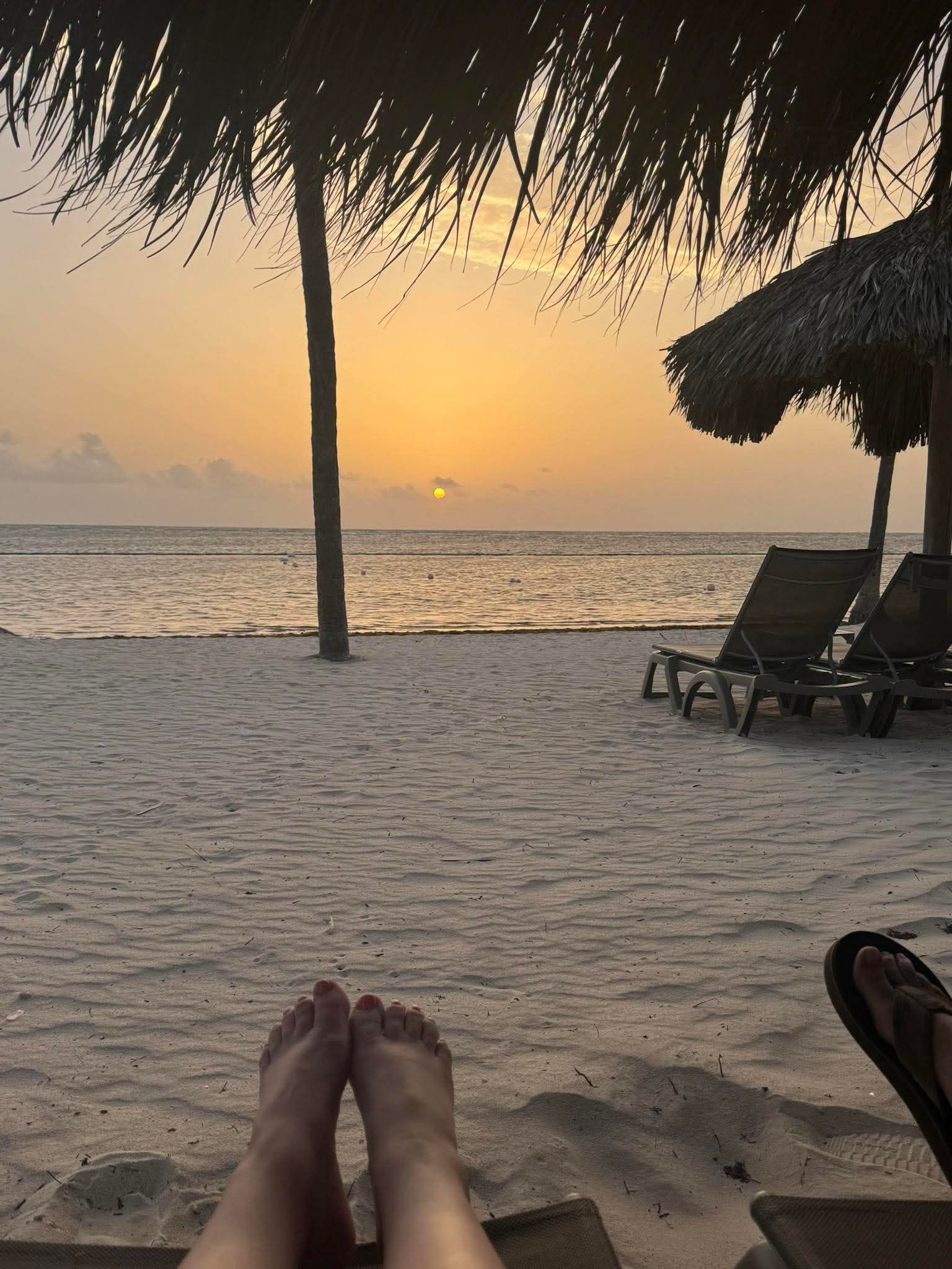 Bare feet rest on a lounge chair overlooking a sandy beach at sunset, with a thatched umbrella and ocean in the distance.