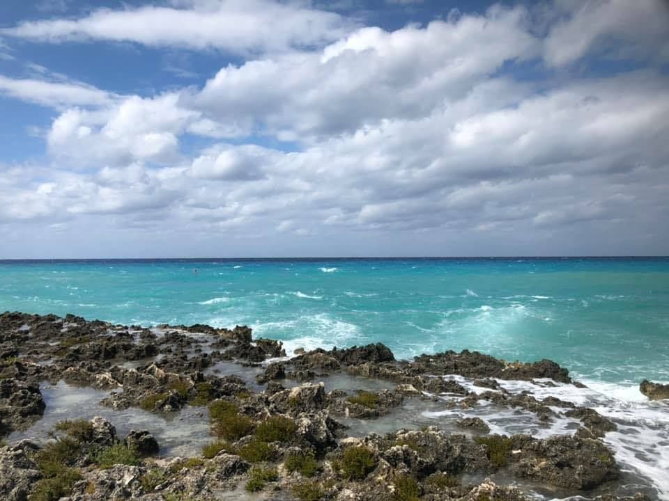 Bright turquoise ocean waves crashing against a rugged, dark rocky shoreline under a cloudy blue sky.