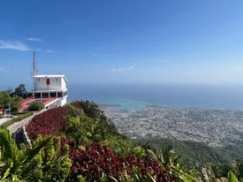 A white hilltop building with a tall antenna overlooking a coastal city and the ocean under a clear blue sky.