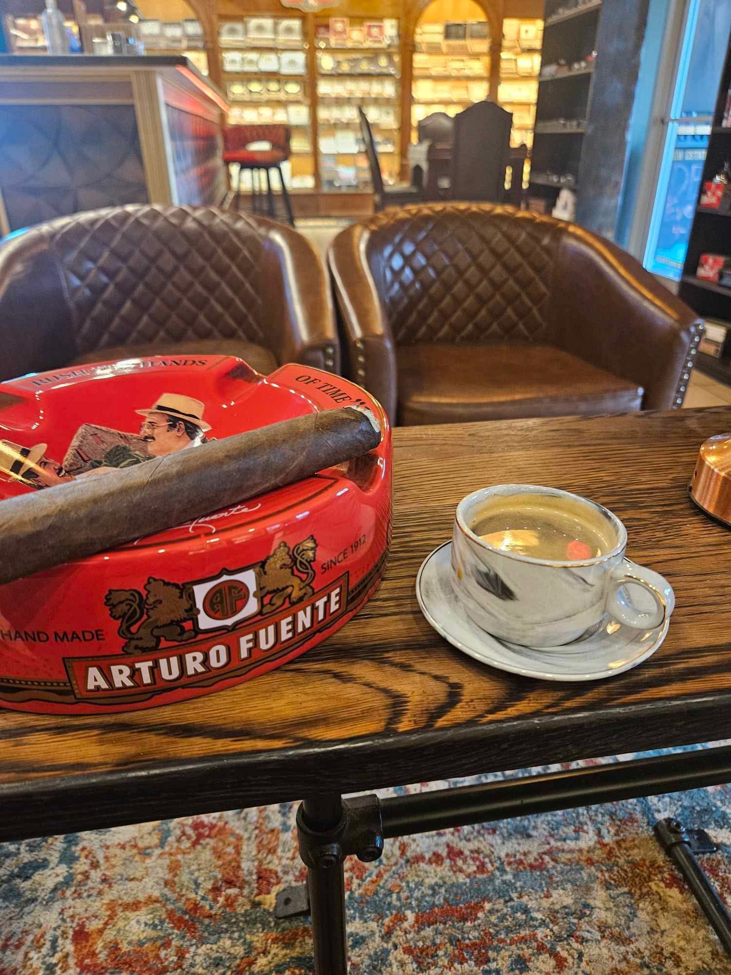 A cigar resting on a red Arturo Fuente ashtray next to a tea cup on a wooden table in a lounge with leather chairs.
