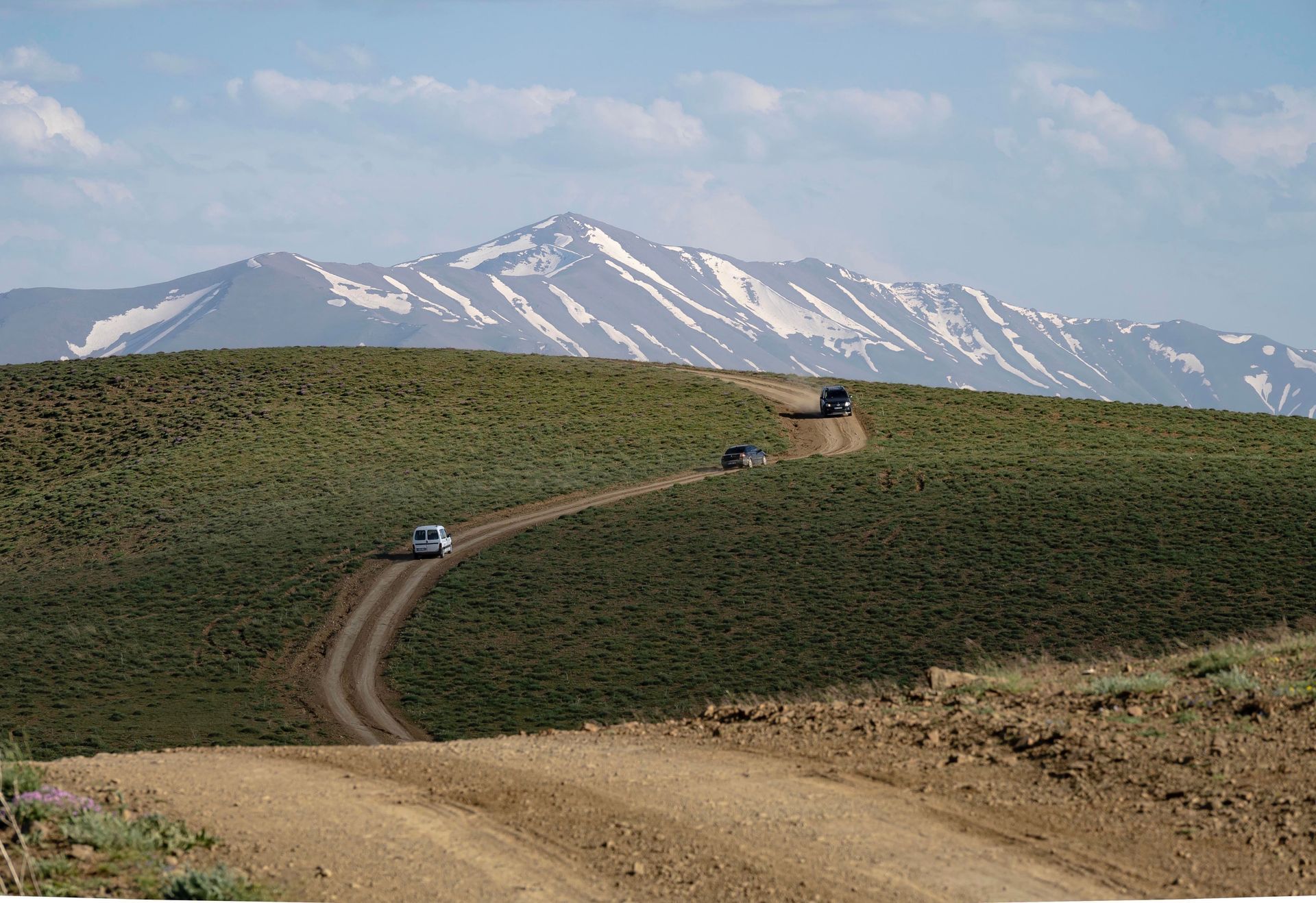Dirt road winding through green hills with a few cars, snow-capped mountains in the background
