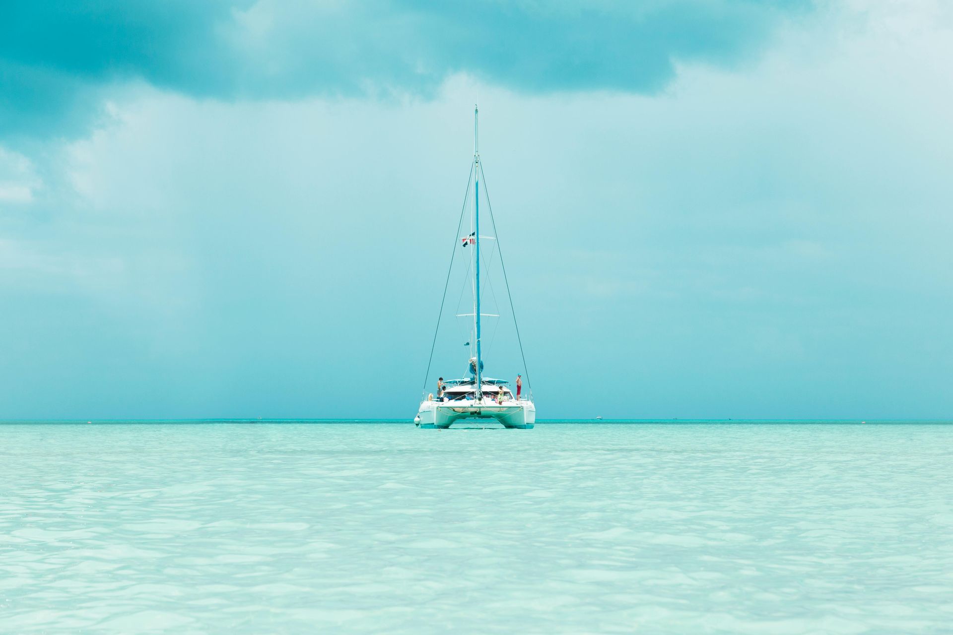 A catamaran sailing on calm, turquoise water under a cloudy, teal-toned sky.