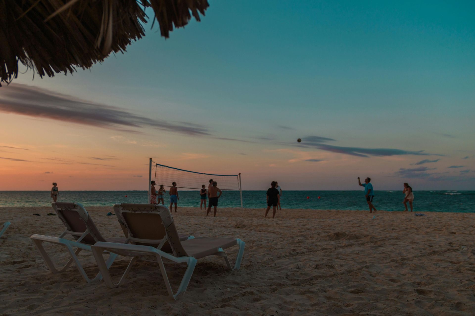 Beach volleyball game at sunset; people playing near the ocean, two lounge chairs in the foreground.